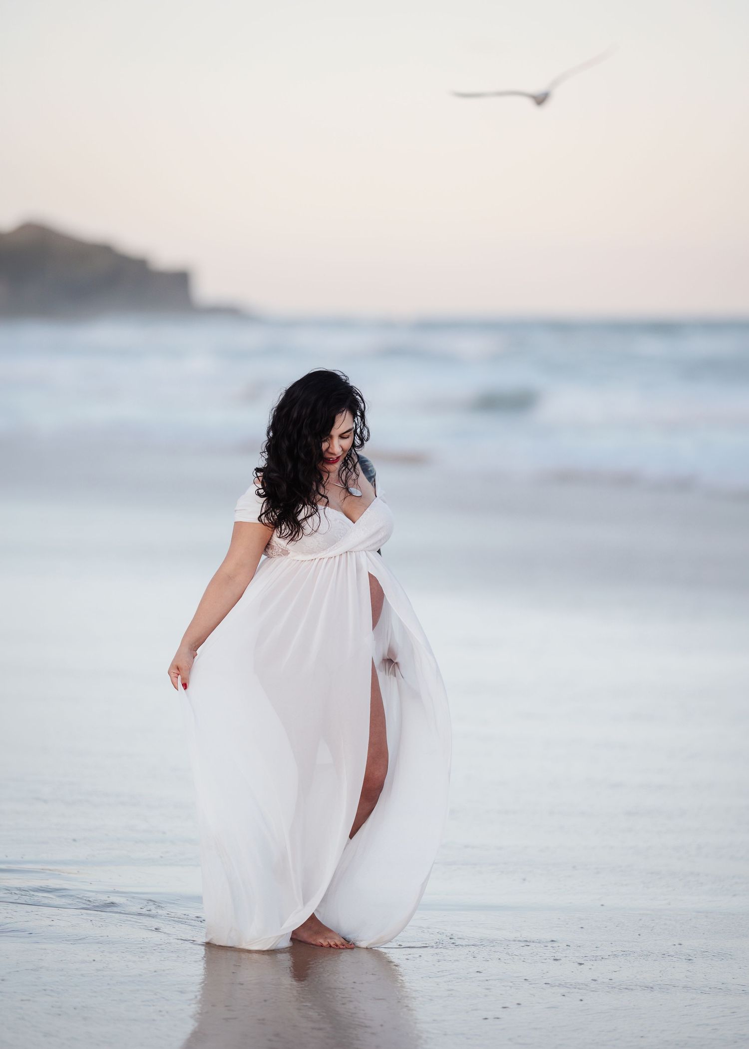 Pregnant woman in flowing white maxi dress walking barefoot on beach at sunset, dark curly hair blowing in breeze.