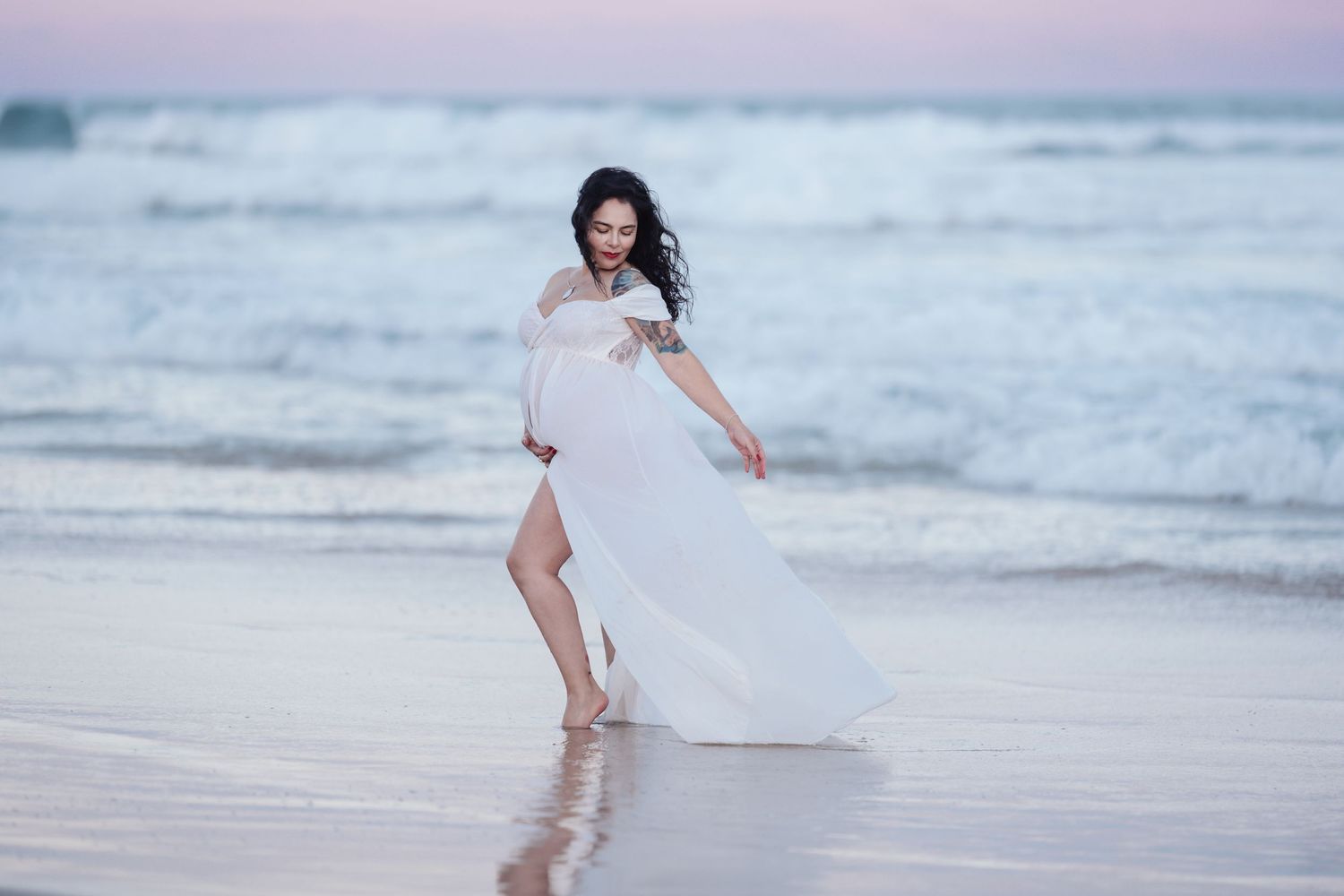 Pregnant woman in white off-shoulder dress posing gracefully on reflective wet beach at sunset.