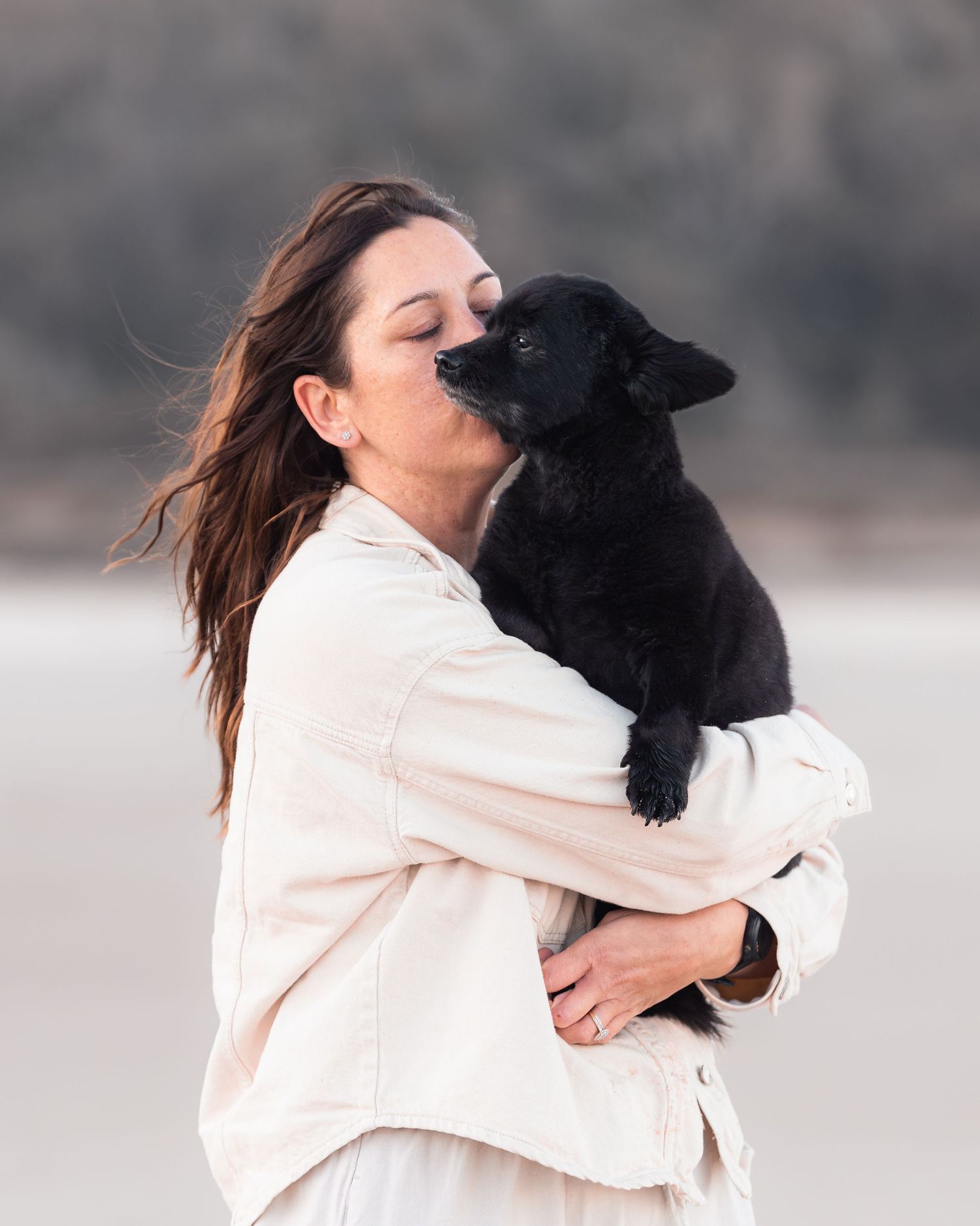 Woman in cream jacket shares a sweet kiss with a small black puppy while standing on a breezy beach.