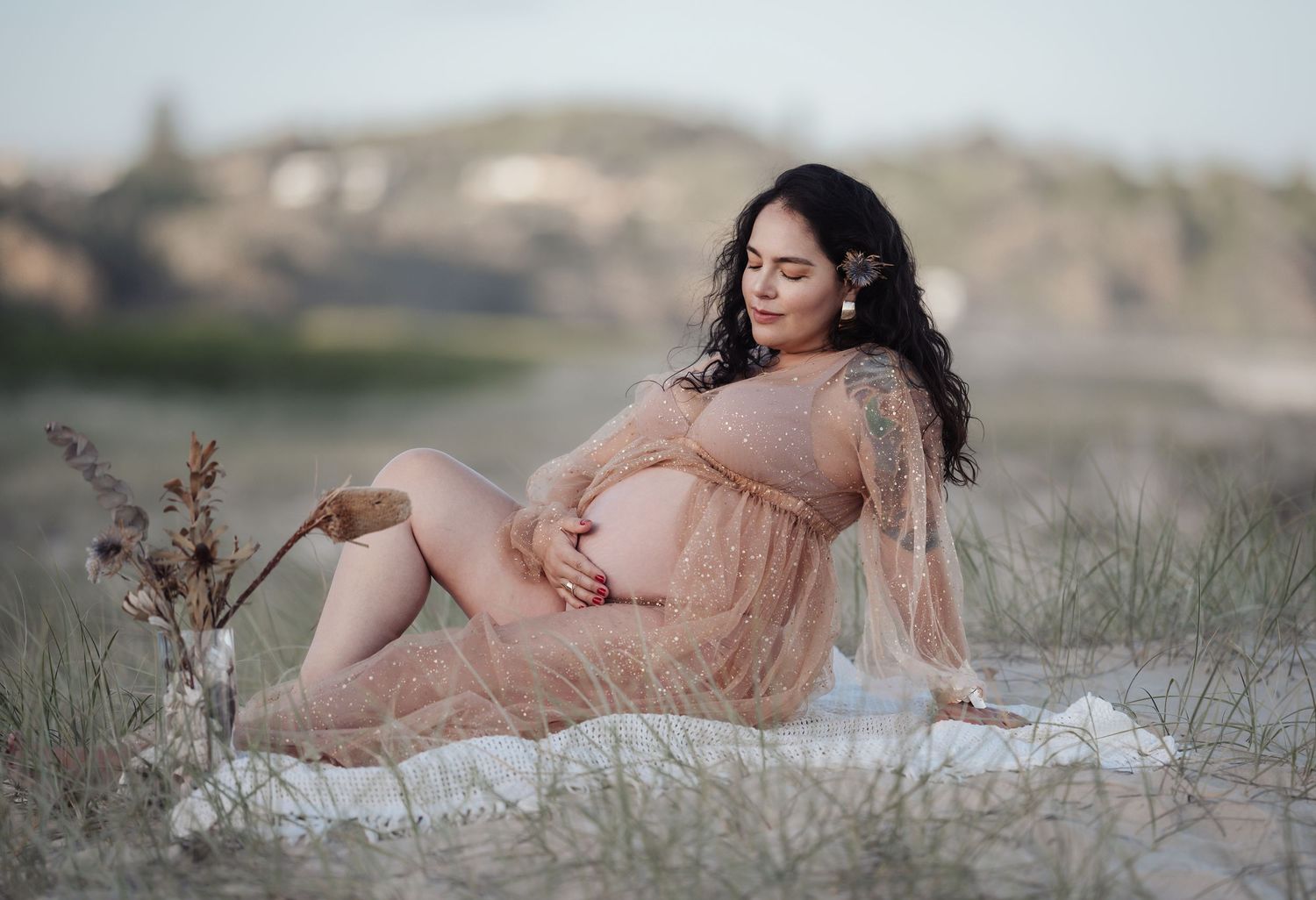 Pregnant woman in sheer peach lace dress reclining outdoors in grassy dunes at golden hour.