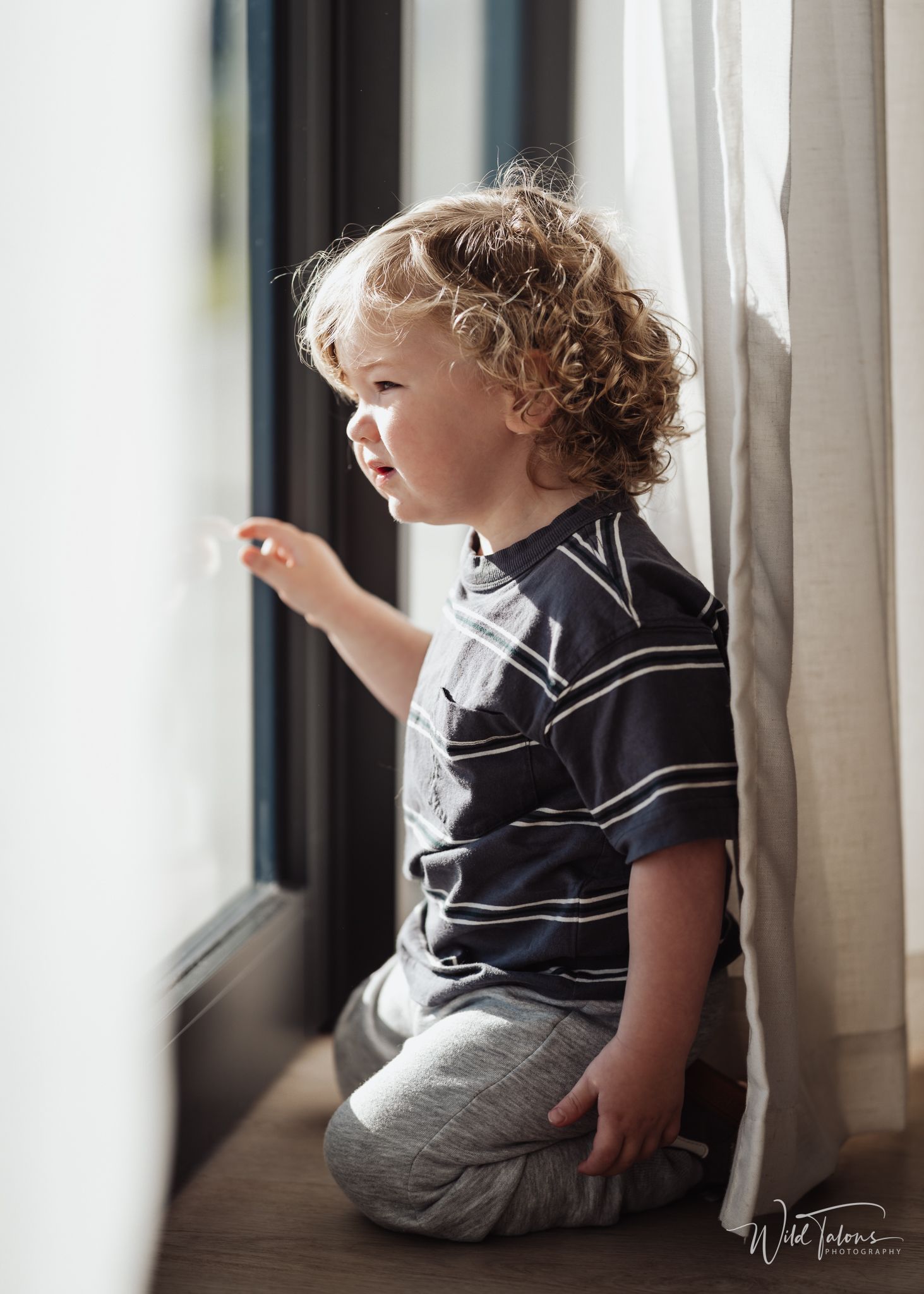 Curly-haired toddler boy in striped shirt kneeling on windowsill, gazing outside through a bright window.