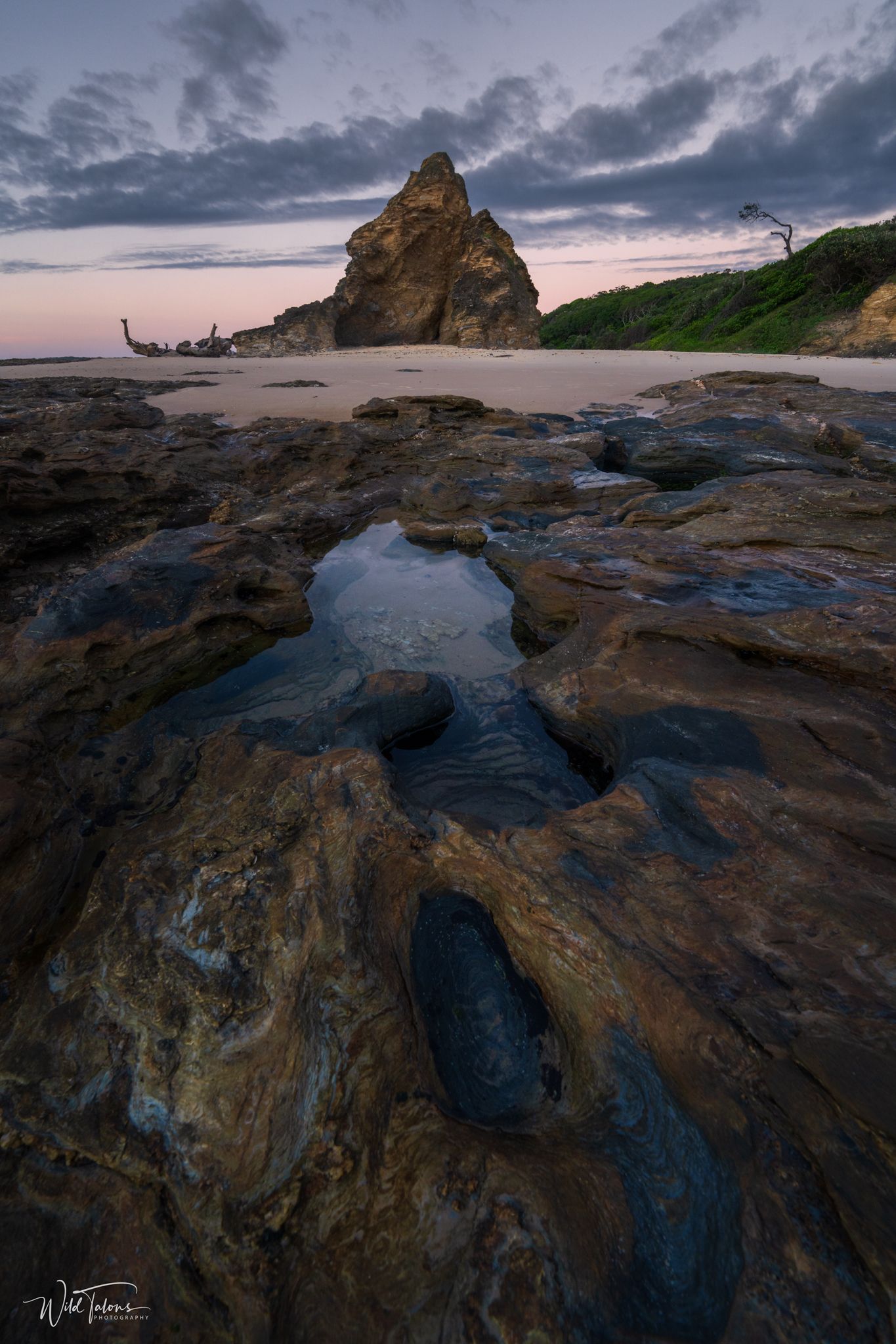 Rugged coastal rock formations with tidal pools lead toward a towering sea stack at twilight on a moody beach.