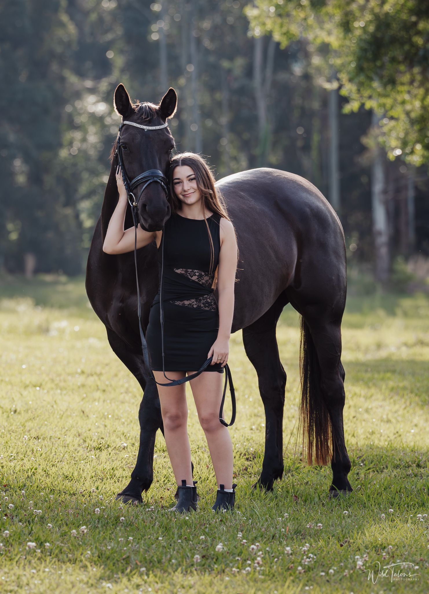 A brunette woman in a black dress poses beside a black horse in a lush green meadow.