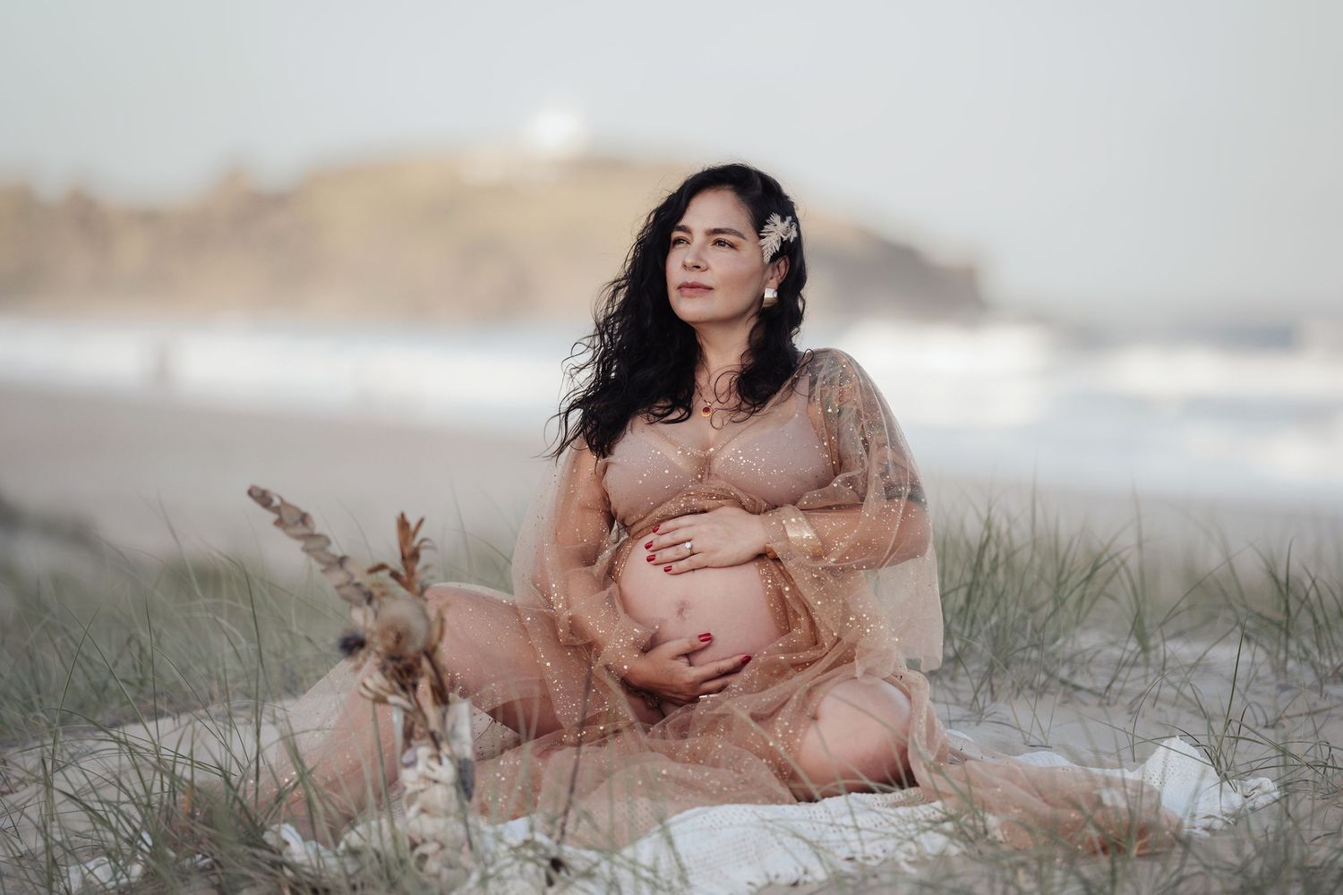 Pregnant woman in sheer beige gown sitting on beach dunes with flower in hair at dusk.