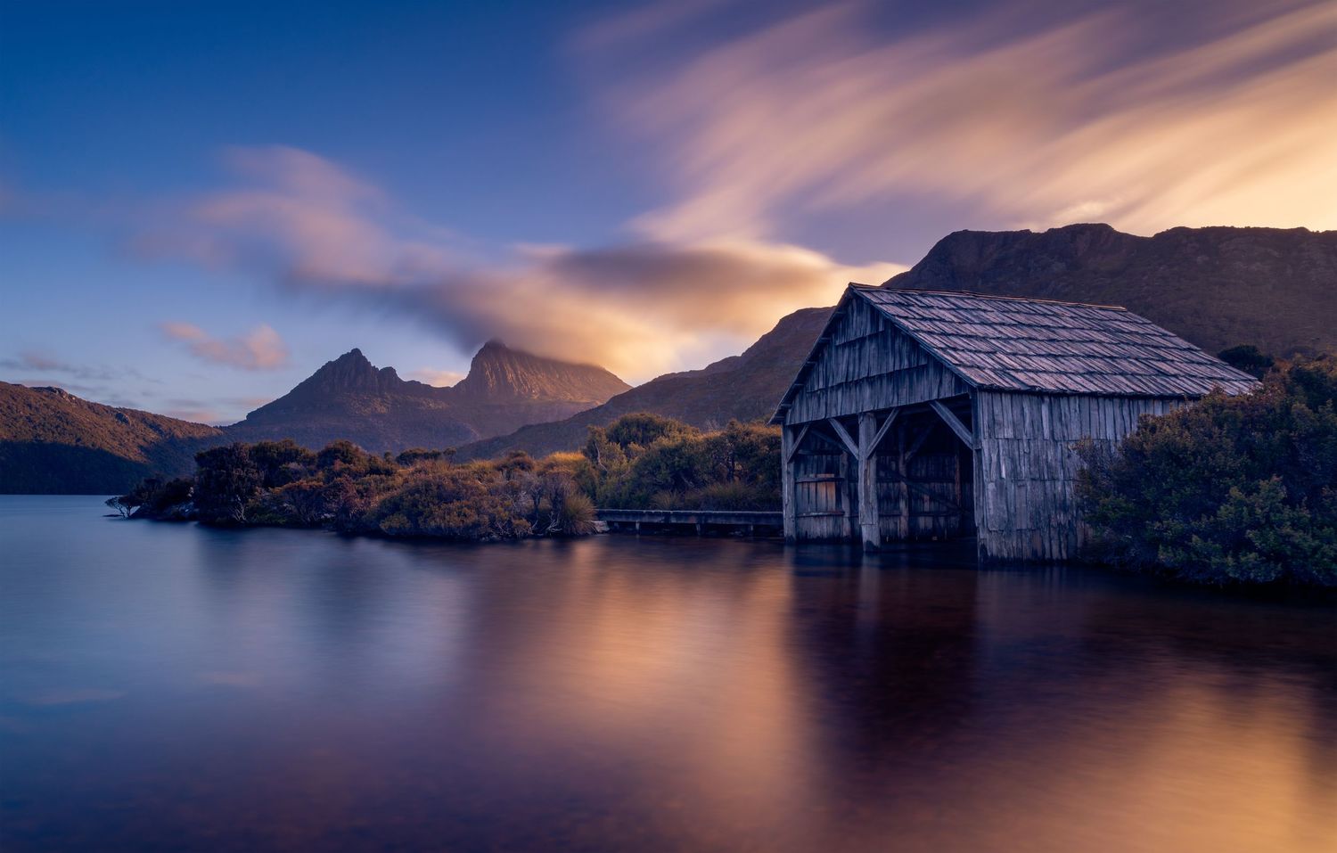 Historic wooden boathouse reflected in calm lake waters at dusk, with Cradle Mountain silhouetted against a glowing sky.