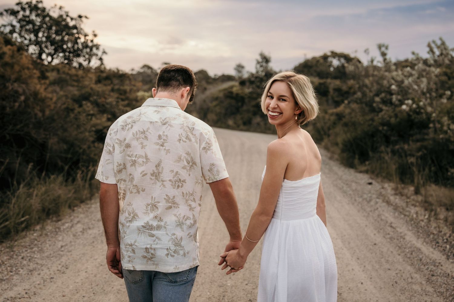 Happy blonde woman in white dress walks hand in hand with partner on dirt road.