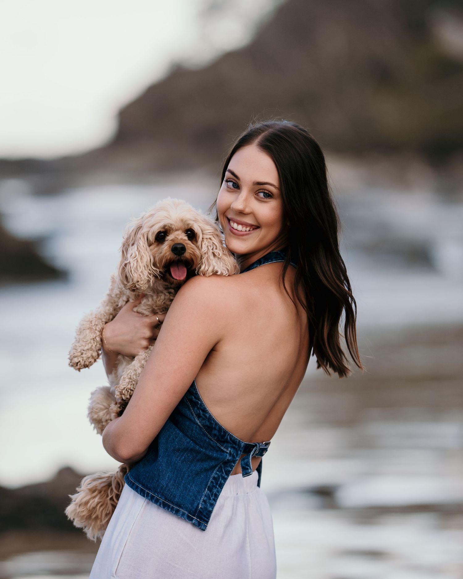 Smiling brunette woman in denim top holding a fluffy cavoodle dog at a rocky beach.