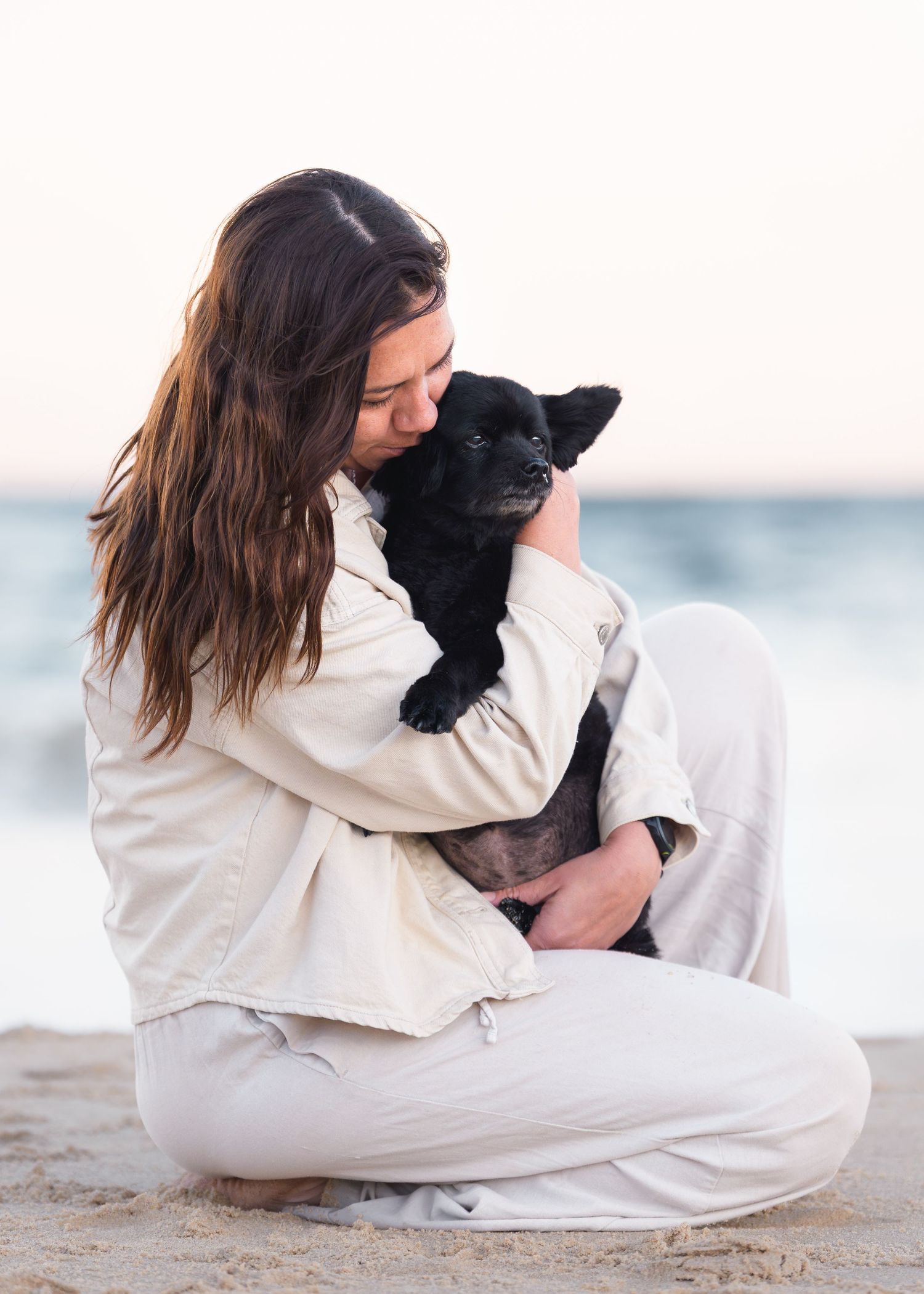 A young woman with long brown hair sits cross-legged on a beach, lovingly cradling a small black puppy.