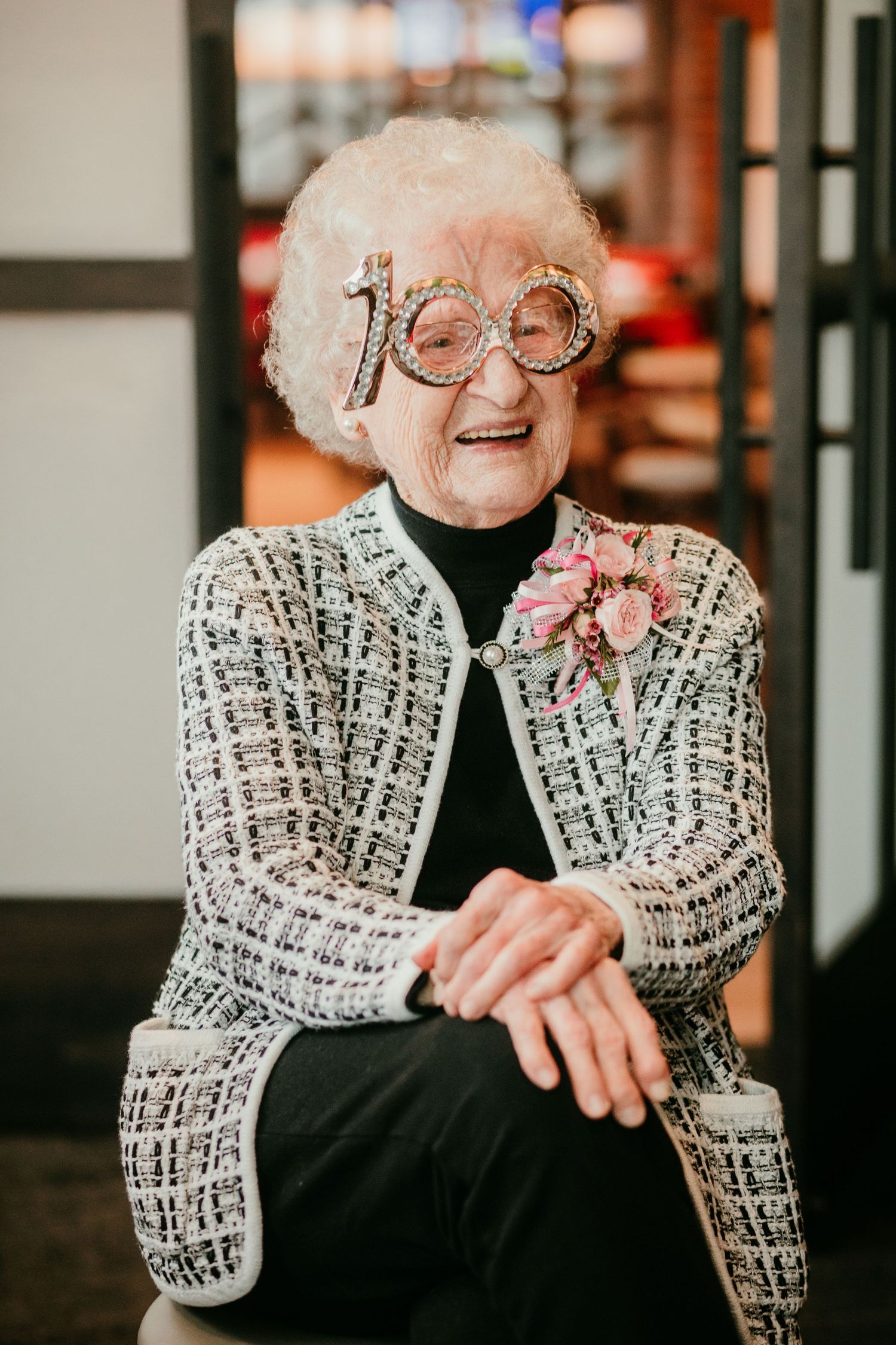 Elderly woman celebrating 100th birthday wearing sparkly number glasses and a floral corsage, smiling joyfully.