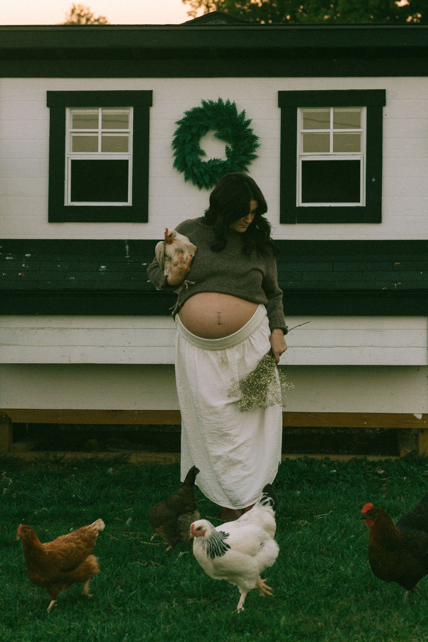 Pregnant woman holding a chicken surrounded by hens outside a white farmhouse chicken coop.