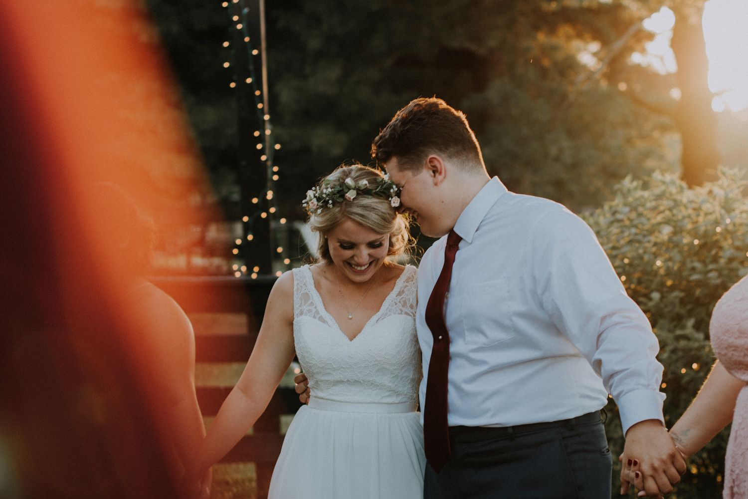 A bride in a floral crown and groom hold hands and smile during a golden hour outdoor wedding ceremony.