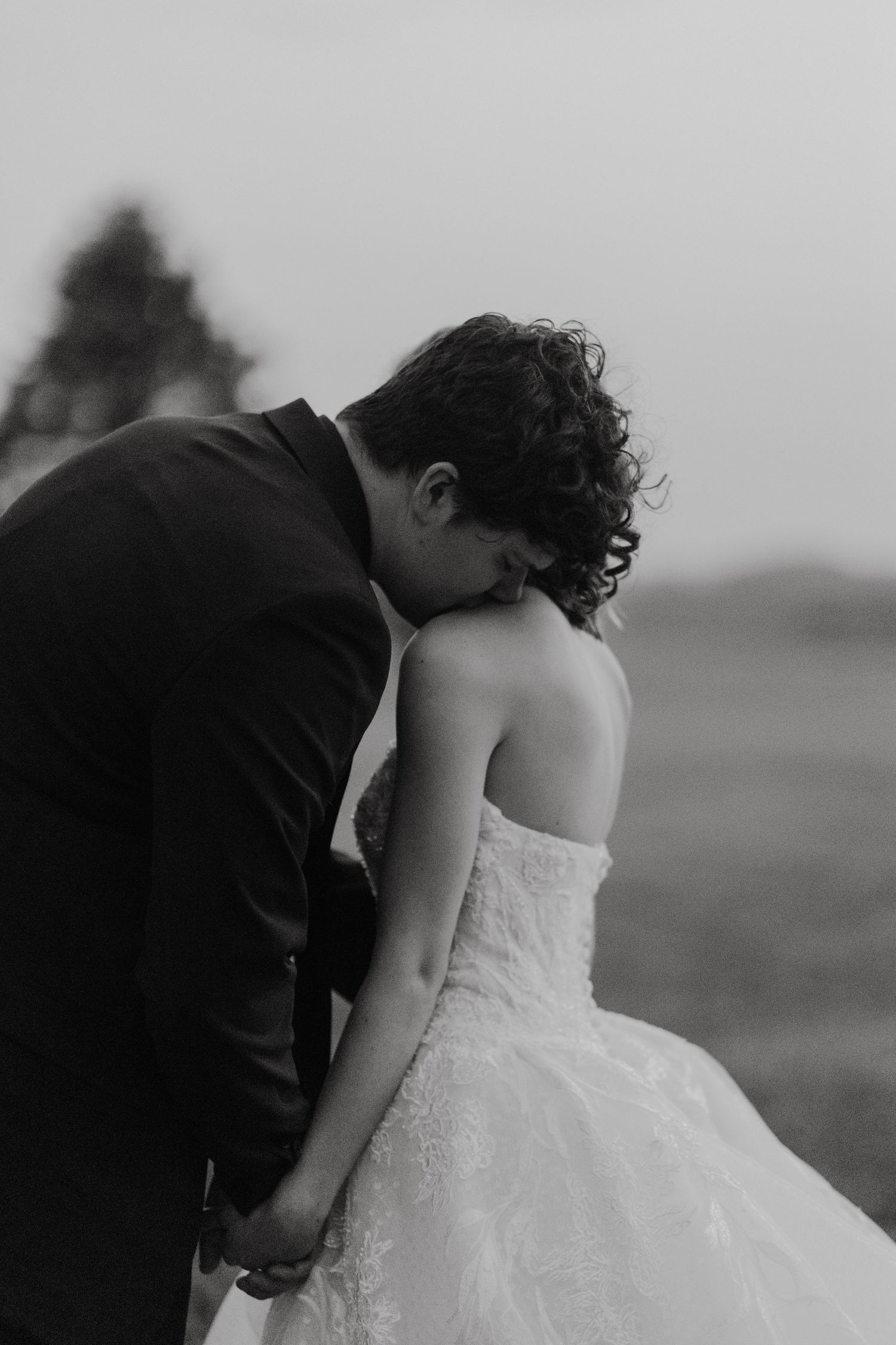 Black and white photo of bride and groom holding hands, groom kissing bride on head outdoors.