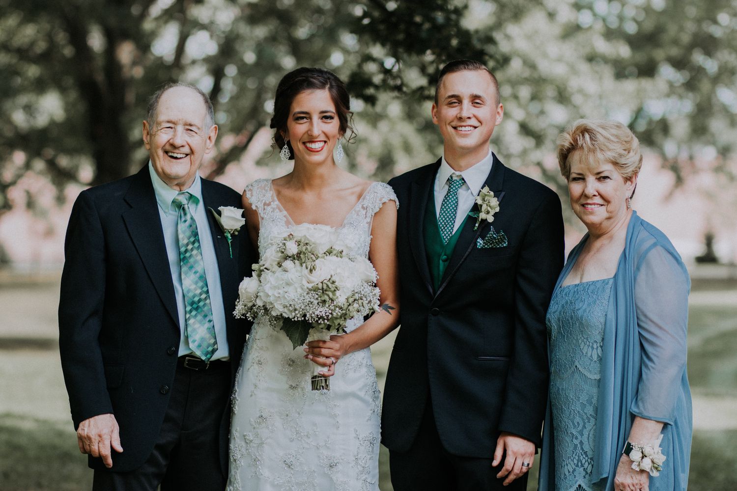 A smiling bride and groom pose with two older family members in a sunny outdoor wedding portrait.