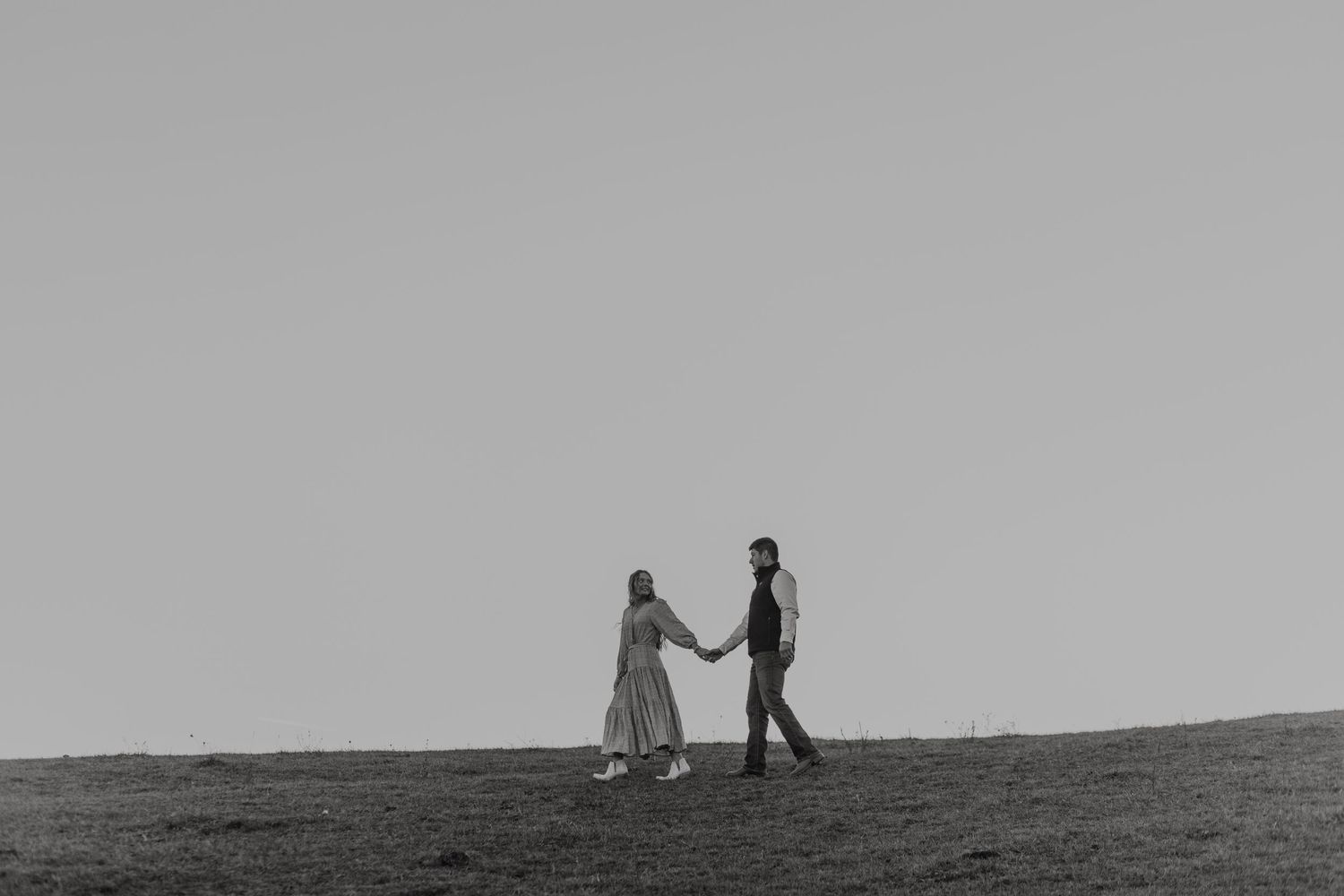 Black and white photo of a couple holding hands while walking across an open grassy hillside.