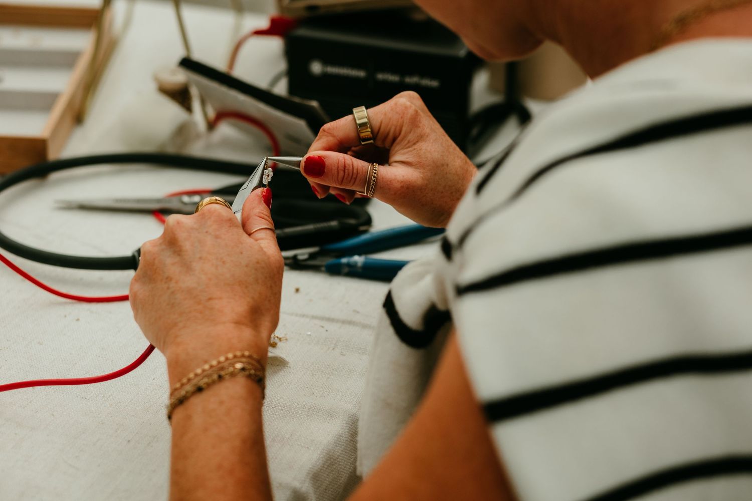 Woman with red nails and gold bracelet soldering jewelry wire at a workbench with tools.
