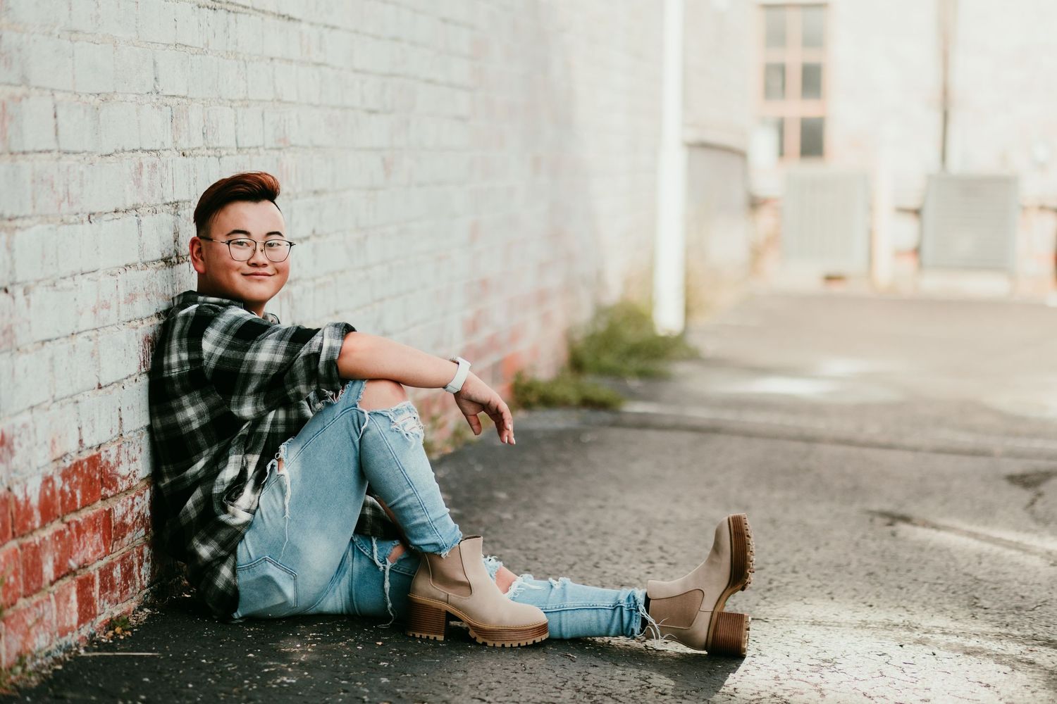 Young person in plaid shirt and jeans sitting against brick wall in alley, smiling, wearing glasses.