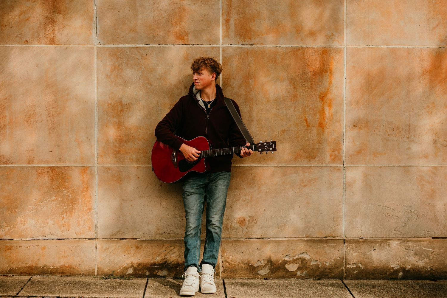 Young guitarist playing red acoustic guitar against a rustic tan stone wall outdoors.