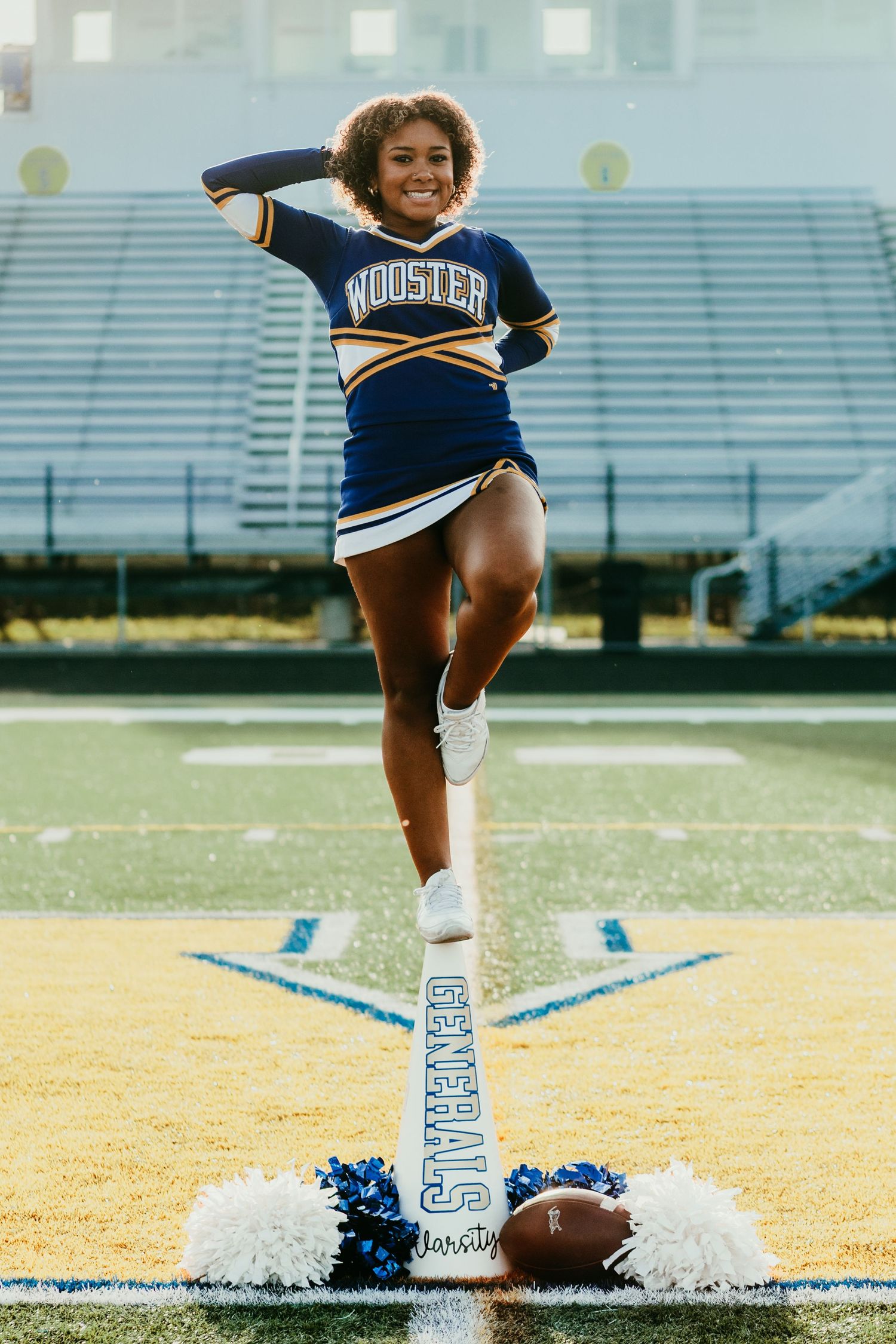 Wooster Generals cheerleader in blue and gold uniform posing on field marker at a football stadium.