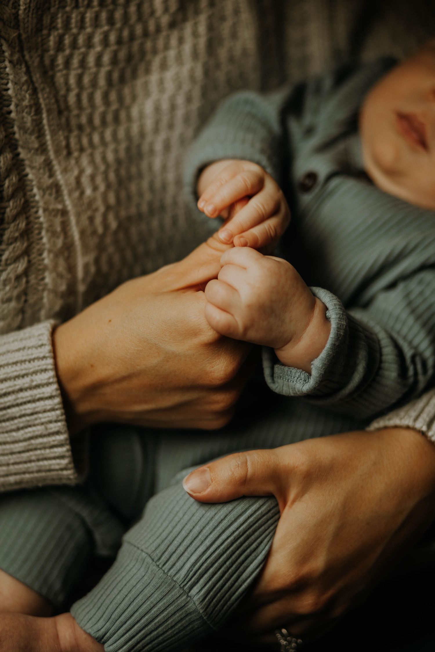 A newborn baby in sage green outfit grasping parent's finger, close-up of tiny hand and adult hands.