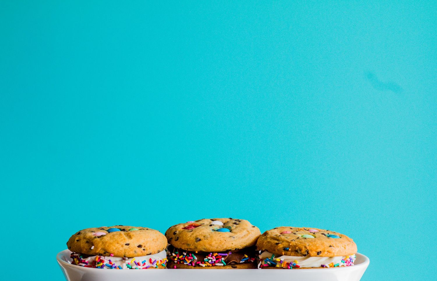 Three colorful sprinkle cookies displayed on a white plate against a bright turquoise background.