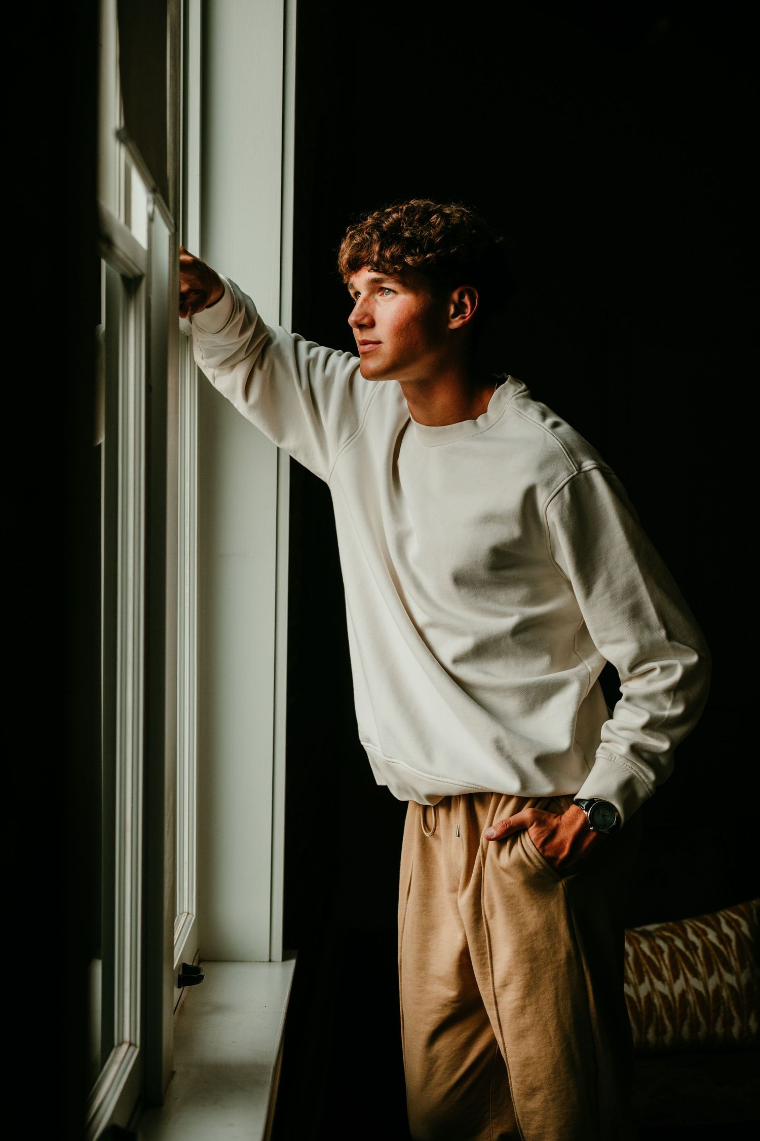 Young man in a white sweatshirt and tan pants gazing out a window in dramatic lighting.