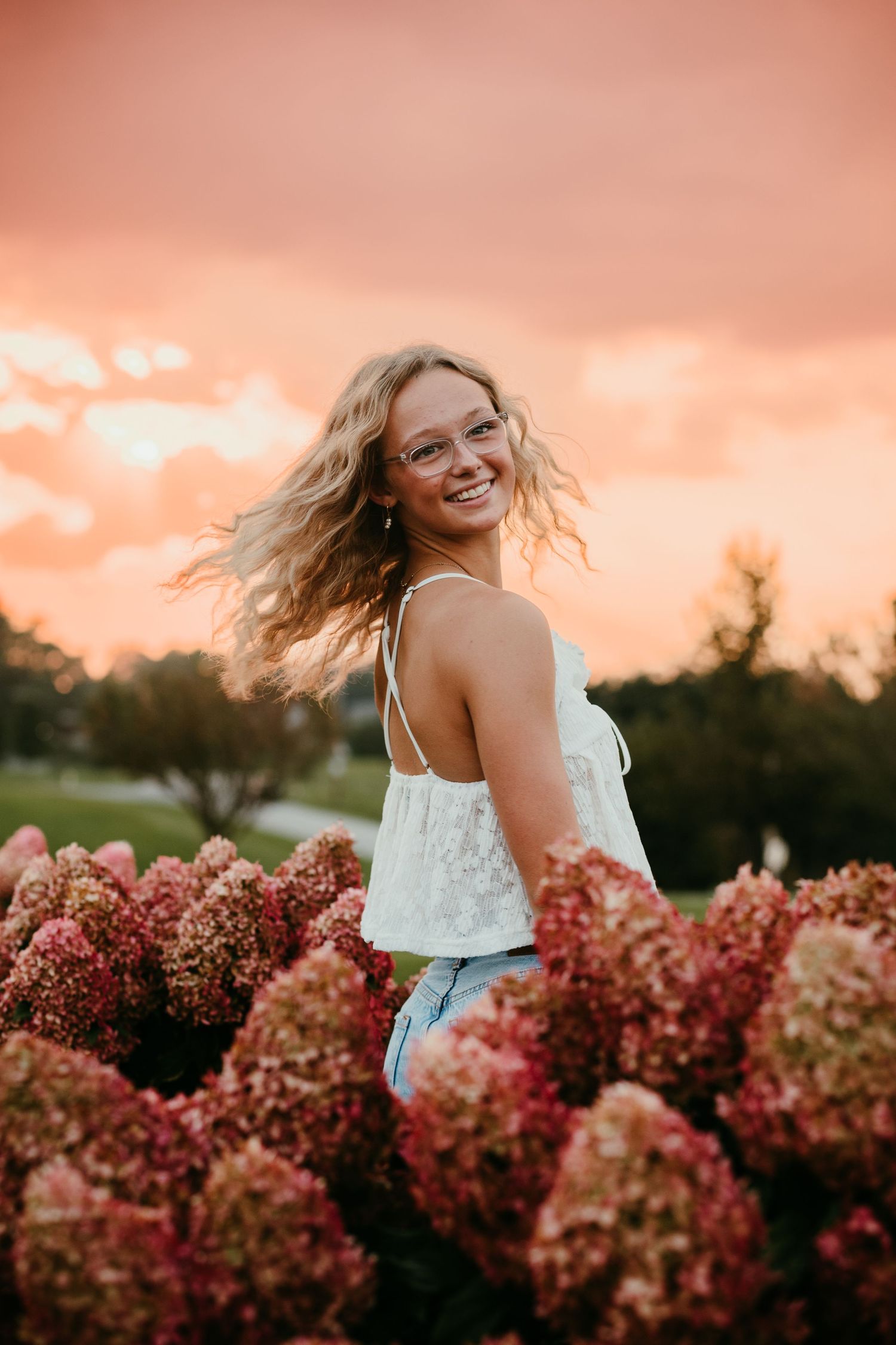 Blonde teenage girl with glasses laughing among pink hydrangeas during a vibrant sunset.