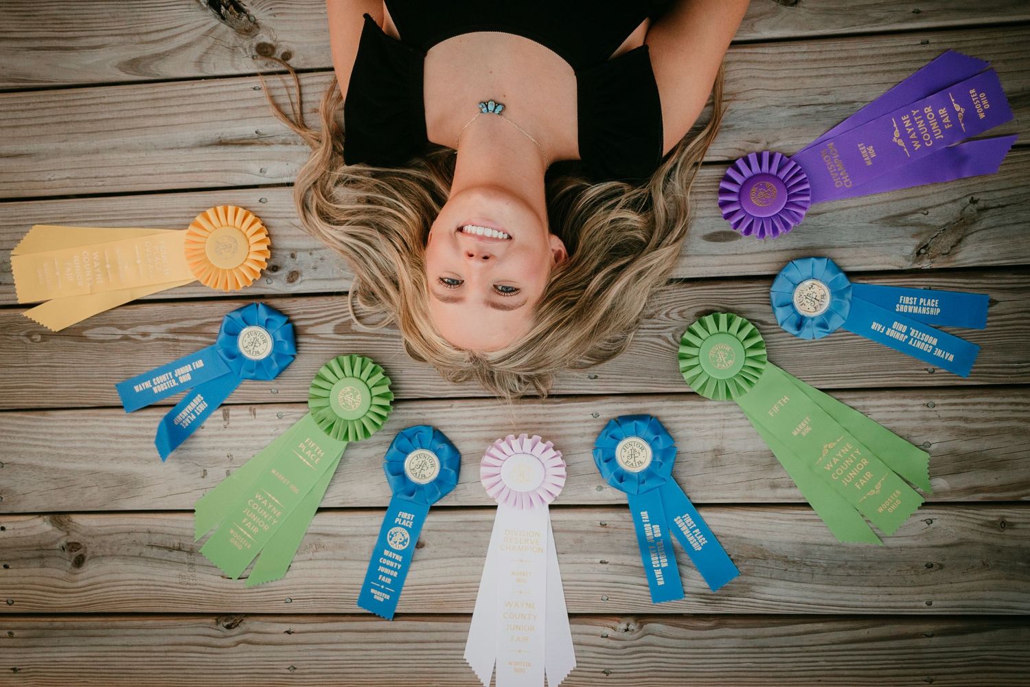 Young woman lying on wooden deck surrounded by colorful award ribbons and competition rosettes.
