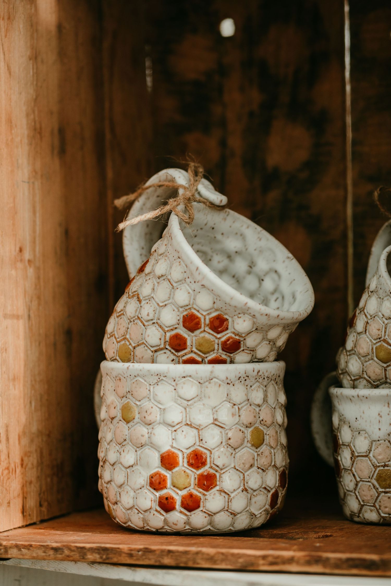 Handmade ceramic honeycomb-patterned mugs stacked on a rustic wooden shelf with twine handles.