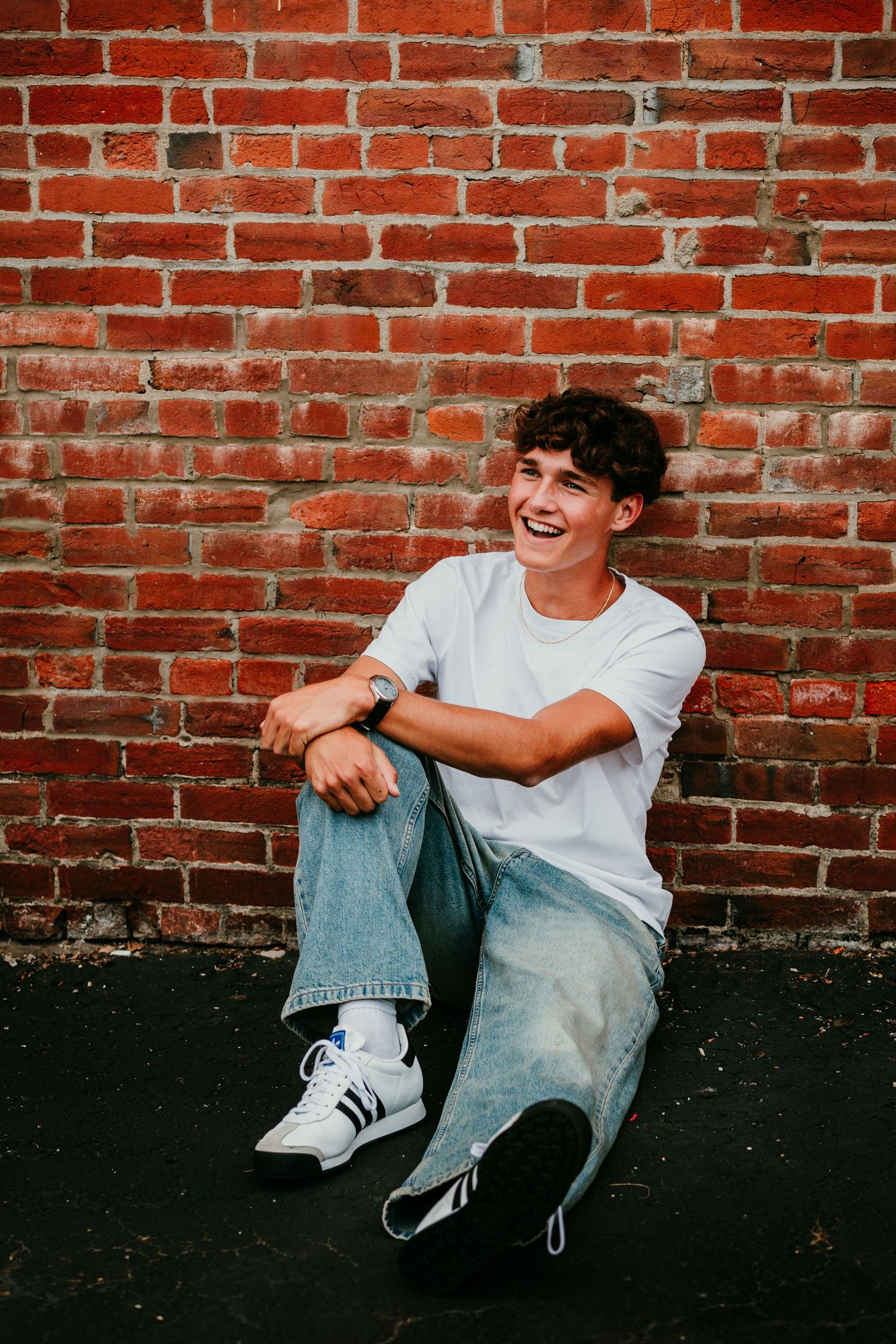Laughing teenage boy sitting against a red brick wall wearing a white t-shirt and jeans.