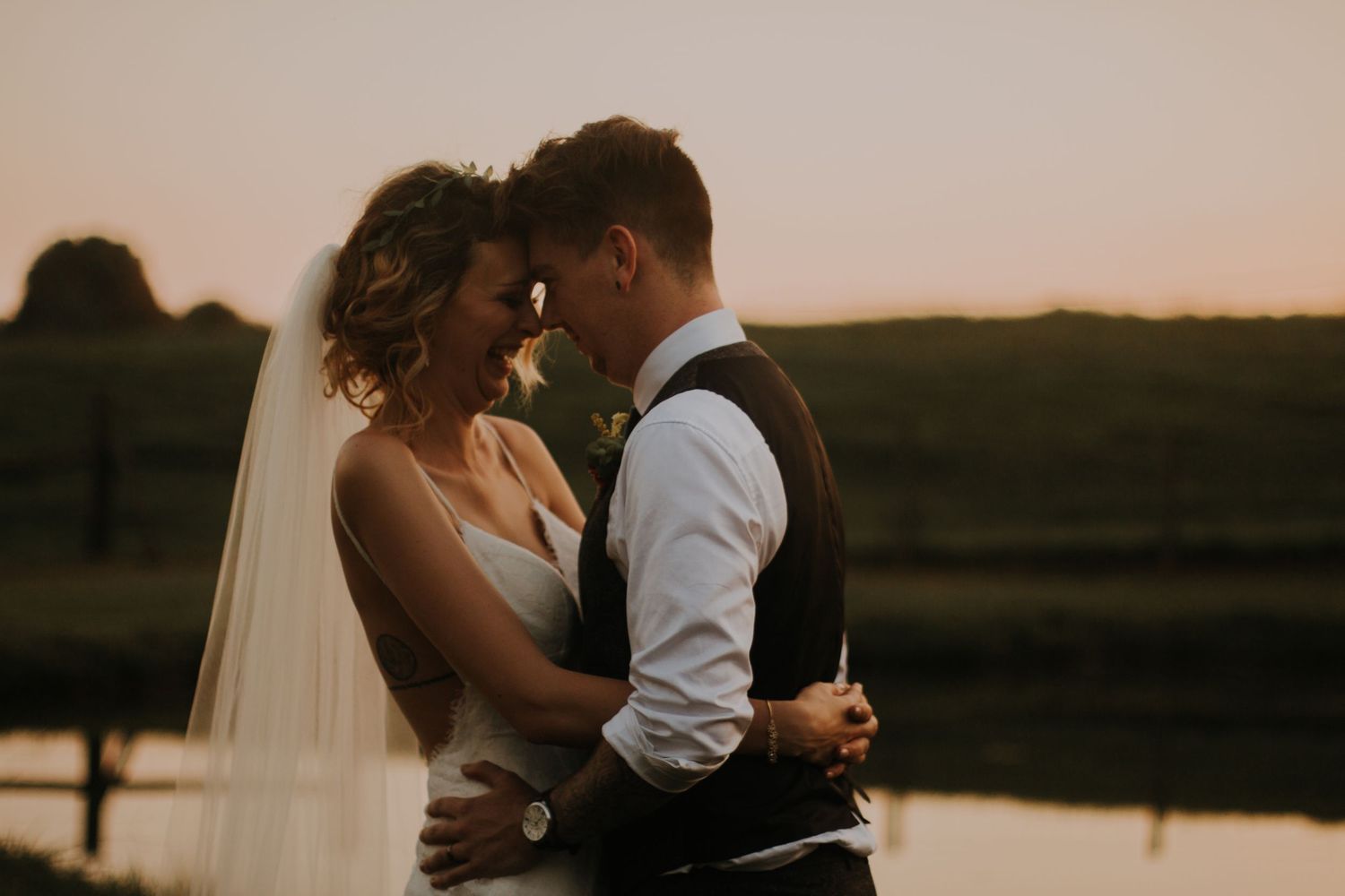 A bride and groom embrace tenderly at golden hour near a peaceful waterfront during their wedding.