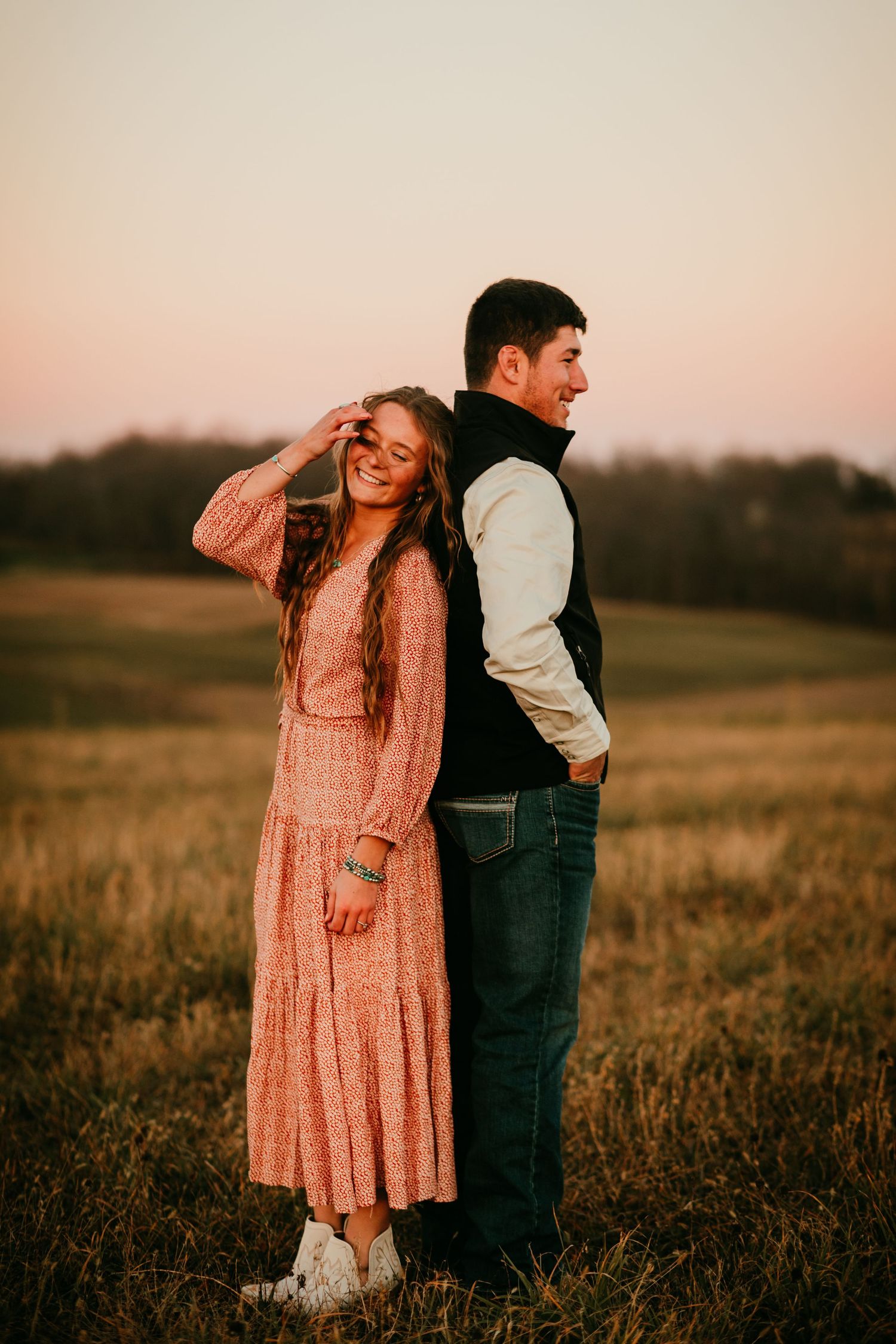 Couple posing at golden hour in a field, woman in pink floral dress smiling over her shoulder.