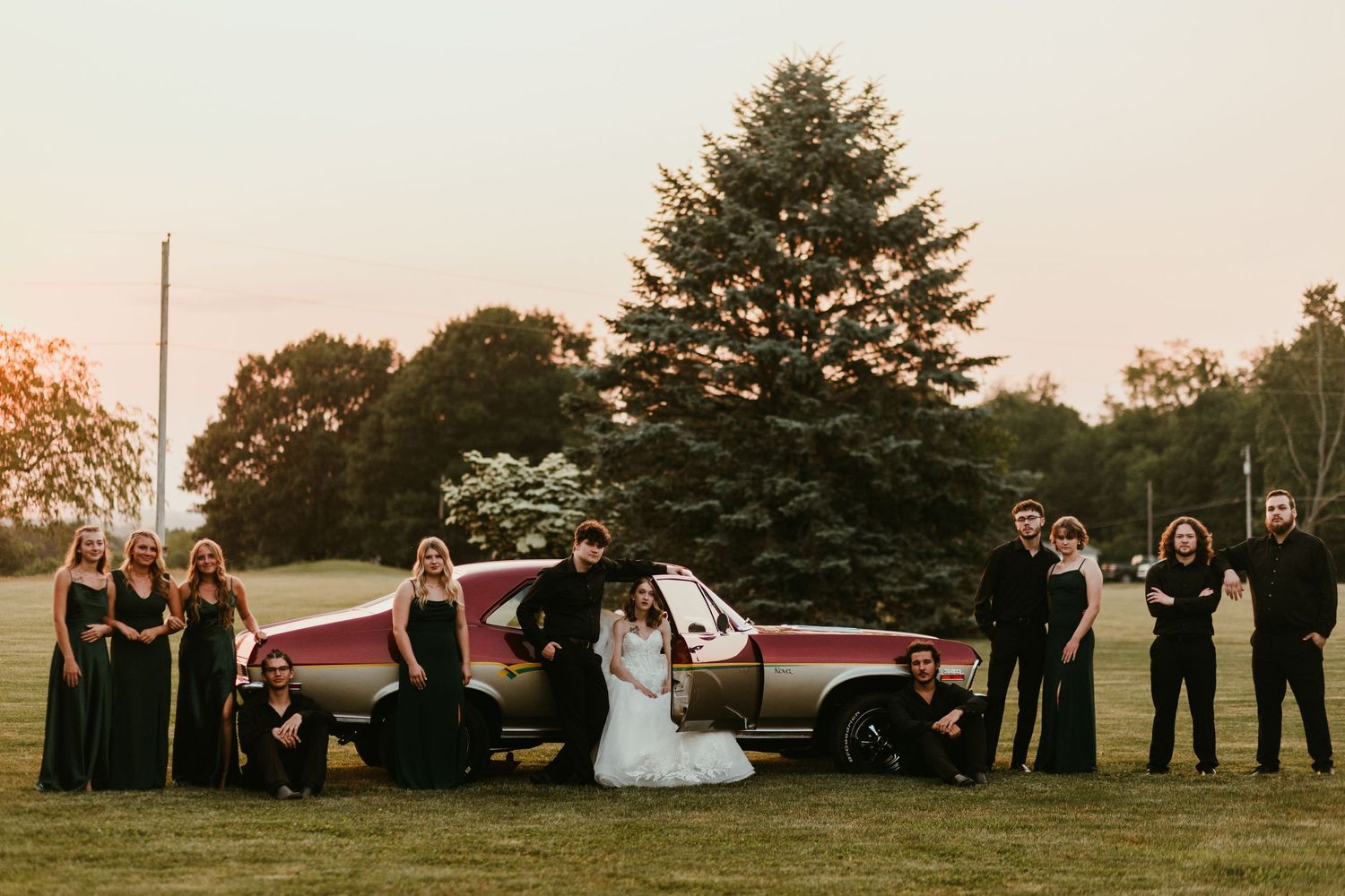 Wedding party posing with a vintage car at sunset beneath a large evergreen tree on a lawn.