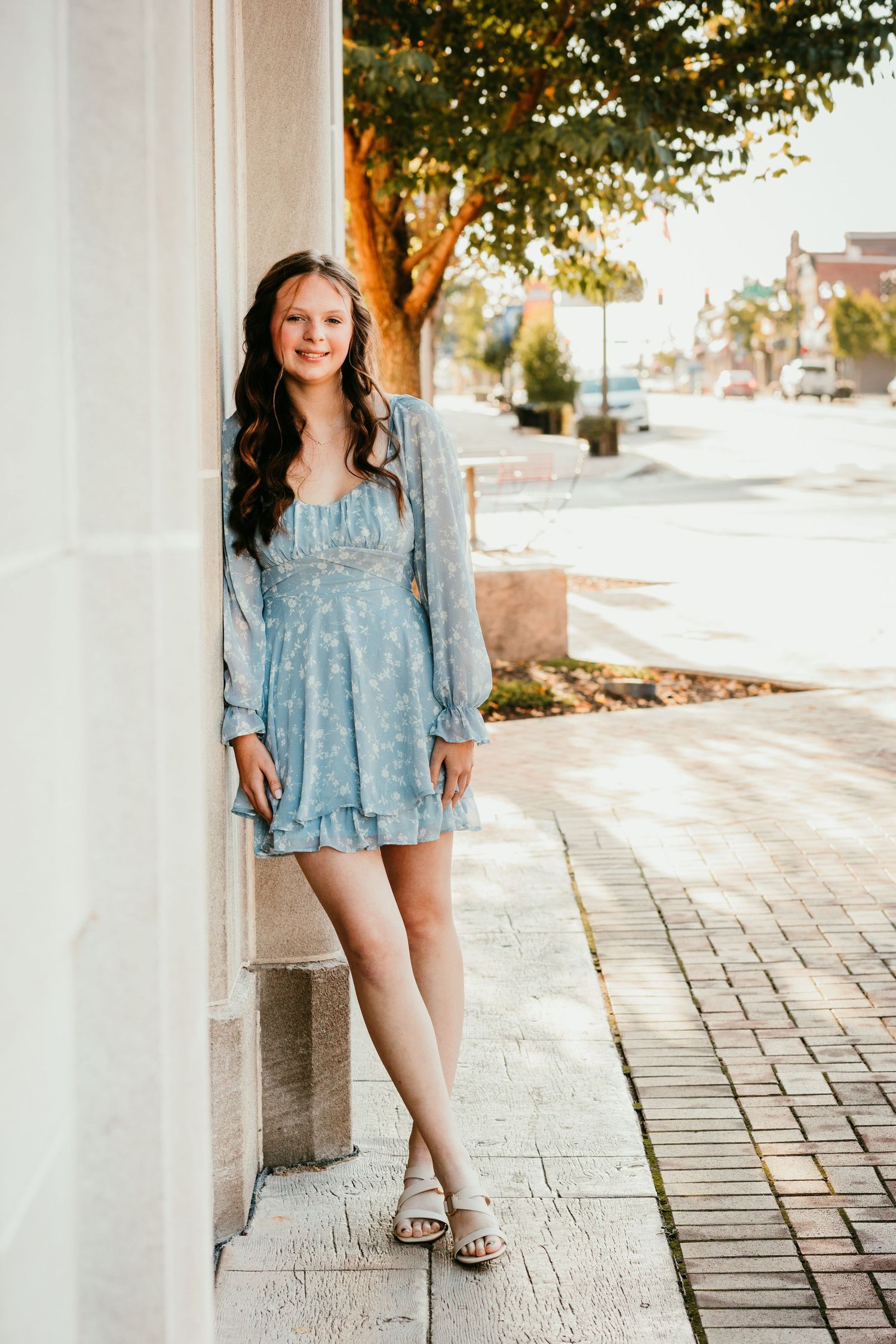 Smiling teenage girl in a light blue long-sleeve mini dress leaning against a column outdoors.
