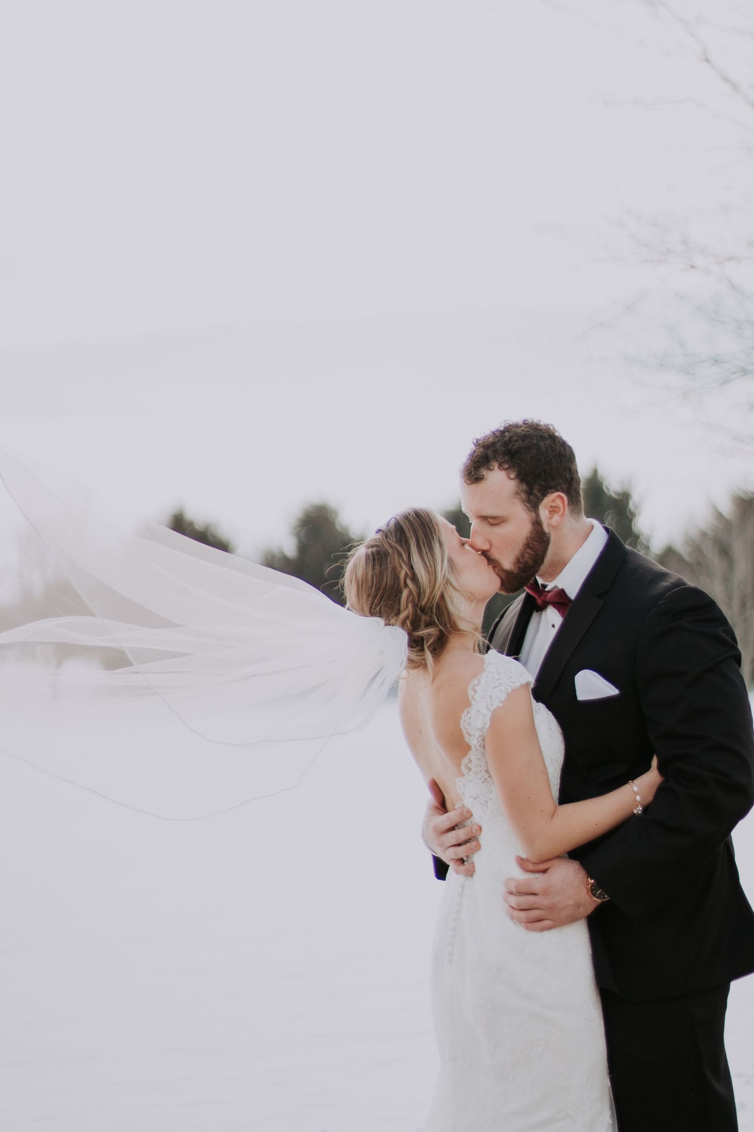 A bride and groom share a romantic kiss as her veil blows in the wind during a winter wedding.