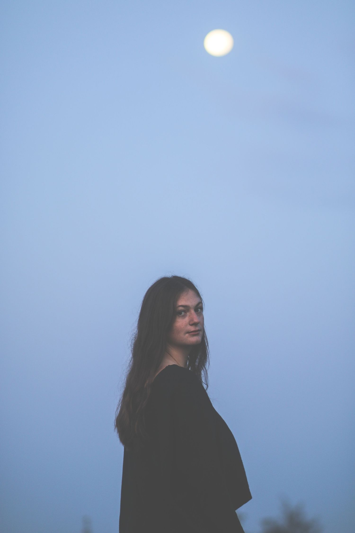 Young woman standing outdoors at dusk with a full moon glowing in the blue sky.