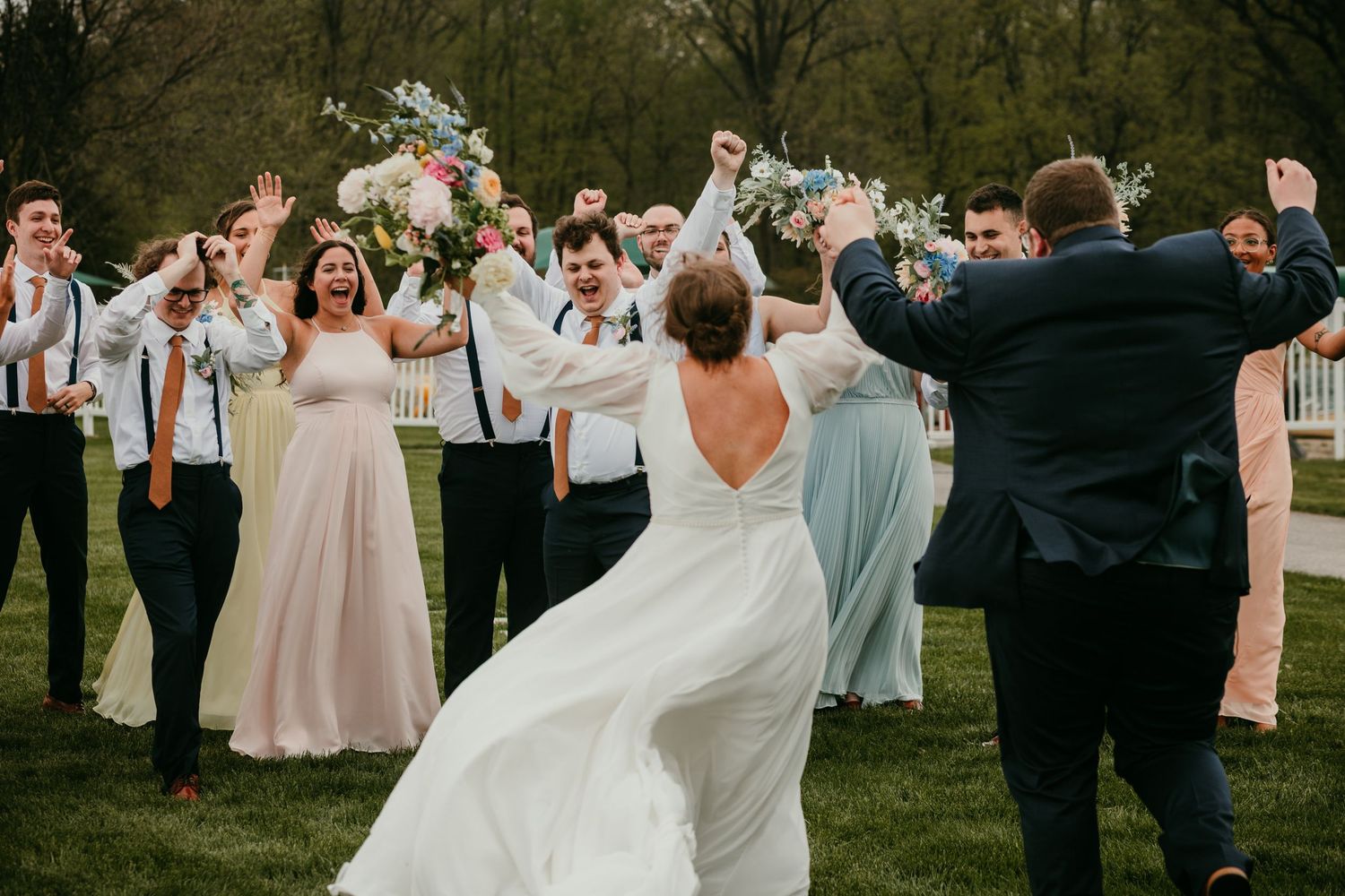 Joyful wedding party celebrating outdoors on a green lawn with bride tossing her floral bouquet.