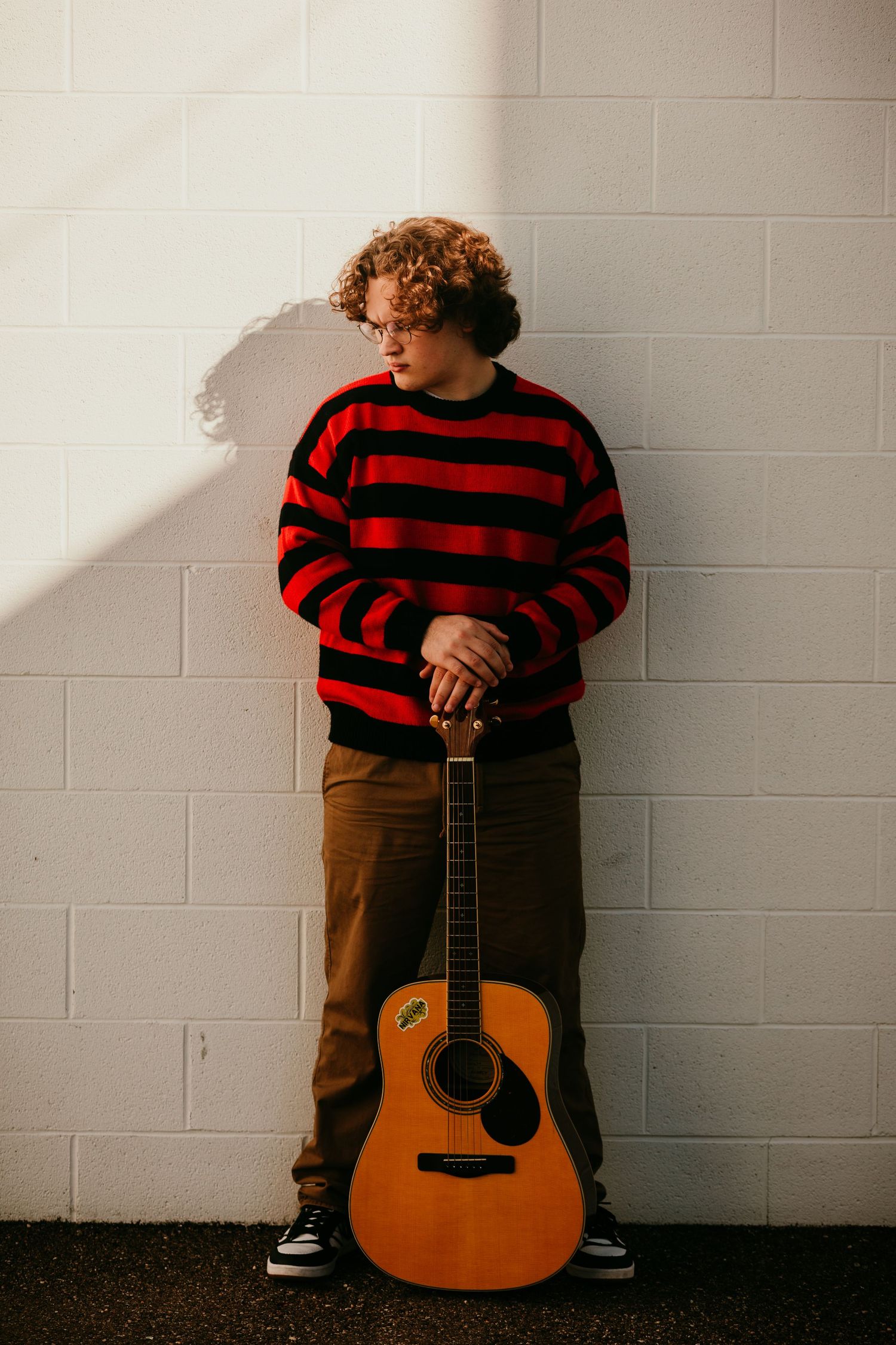 Young curly-haired musician in striped sweater leaning against white brick wall with guitar.