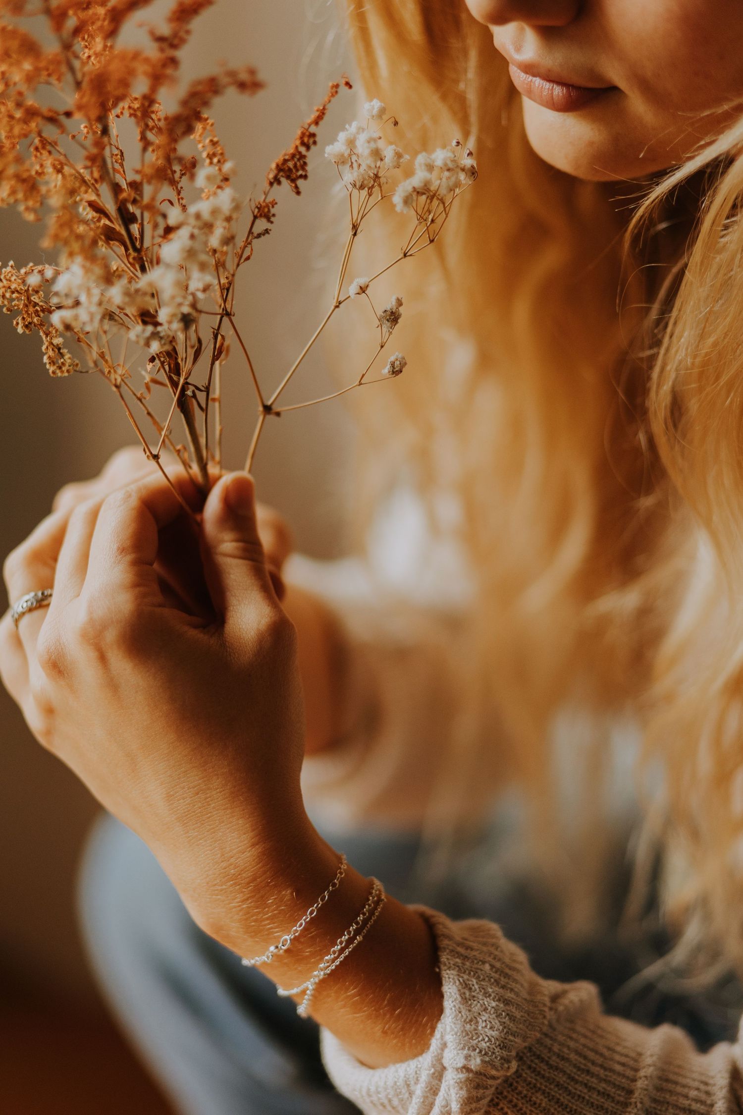 Blonde woman in cozy sweater holding delicate dried wildflowers with silver bracelet.
