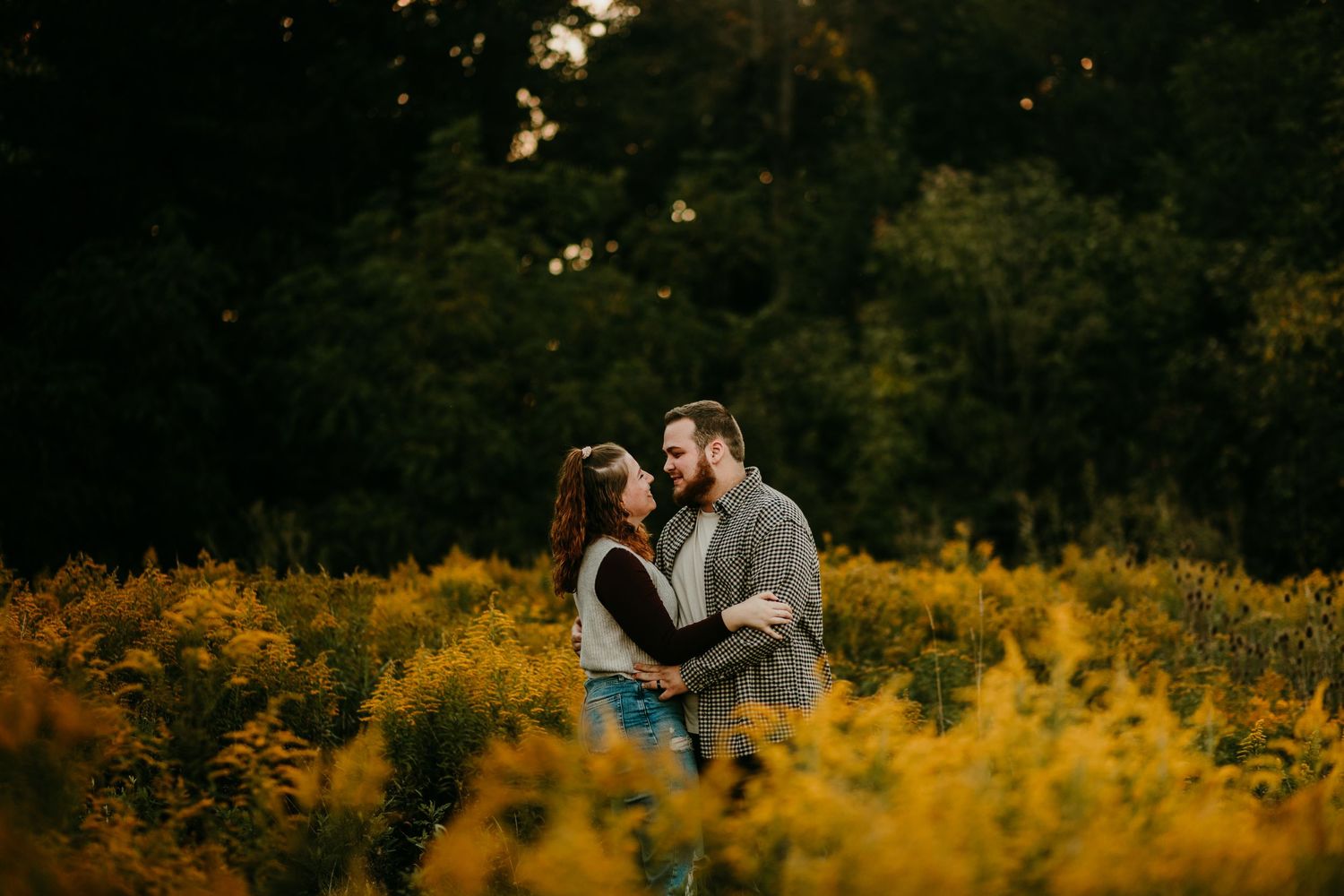 Couple embracing in a golden wildflower field at sunset, surrounded by lush green trees in a romantic outdoor setting.