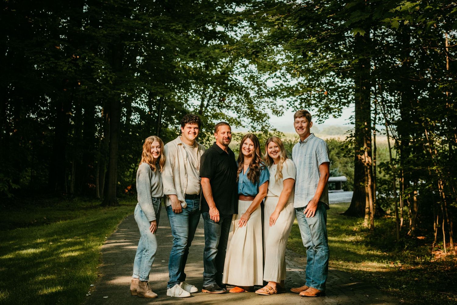 Group of six young adults smiling together on a sunlit tree-lined path outdoors.