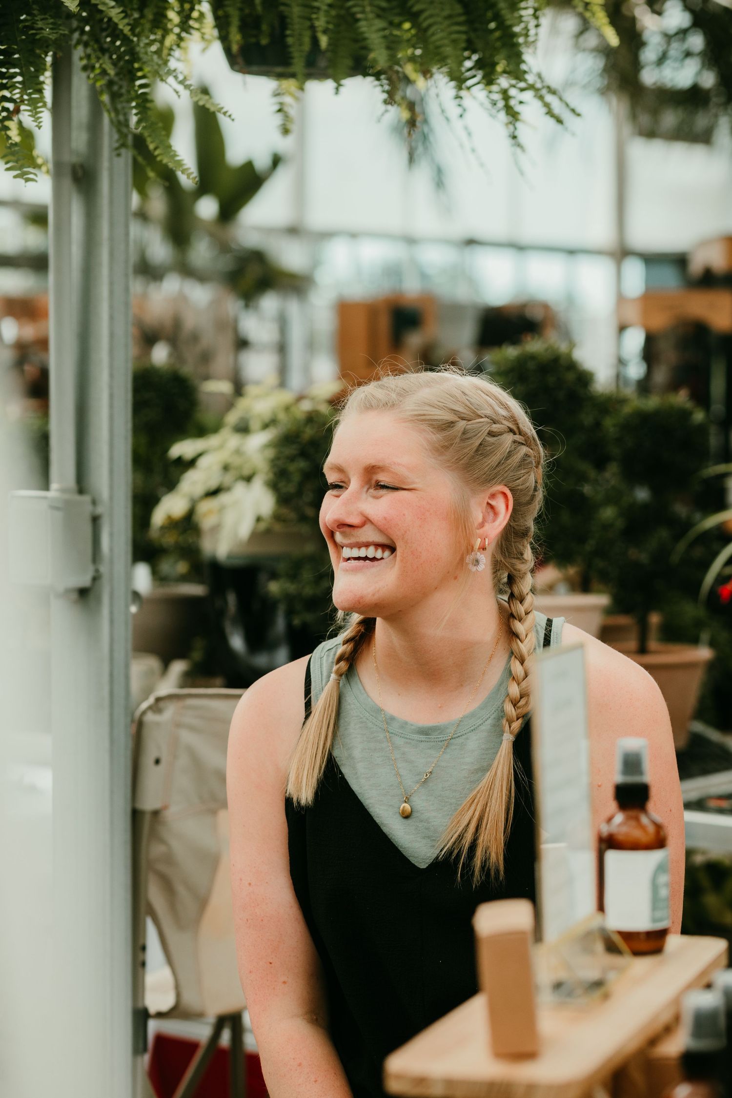 Smiling blonde woman with braids laughing joyfully inside a lush green plant nursery greenhouse.