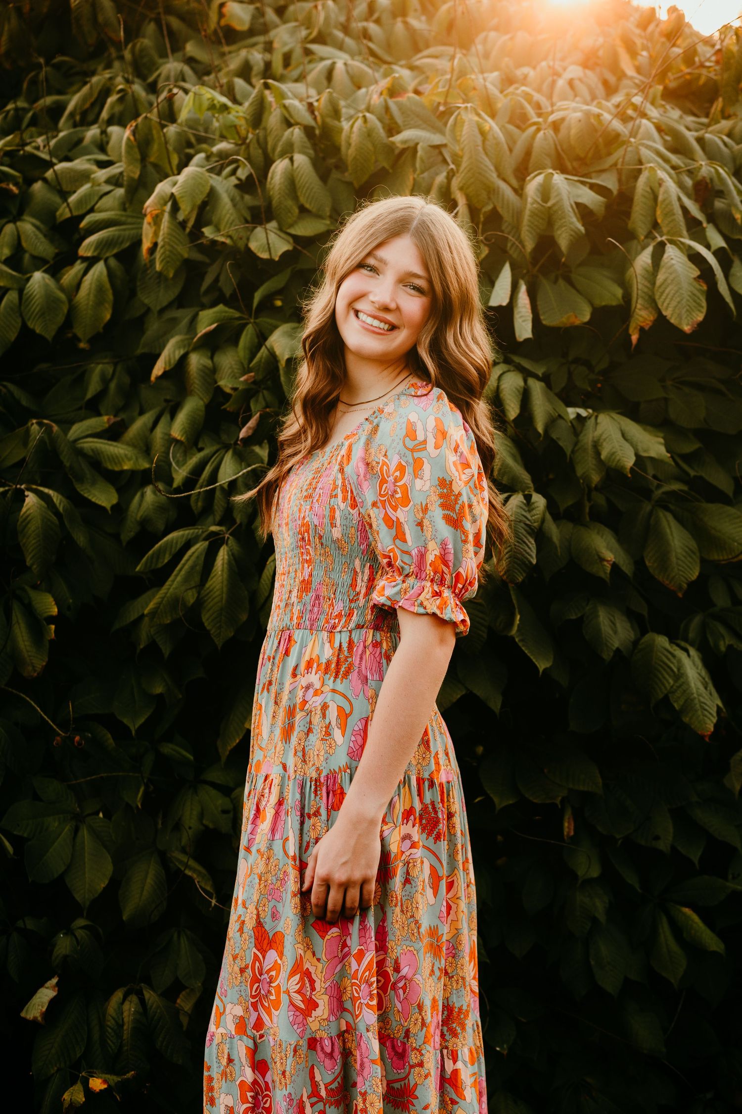 Smiling teenage girl in a colorful floral dress standing in front of lush green foliage at golden hour.