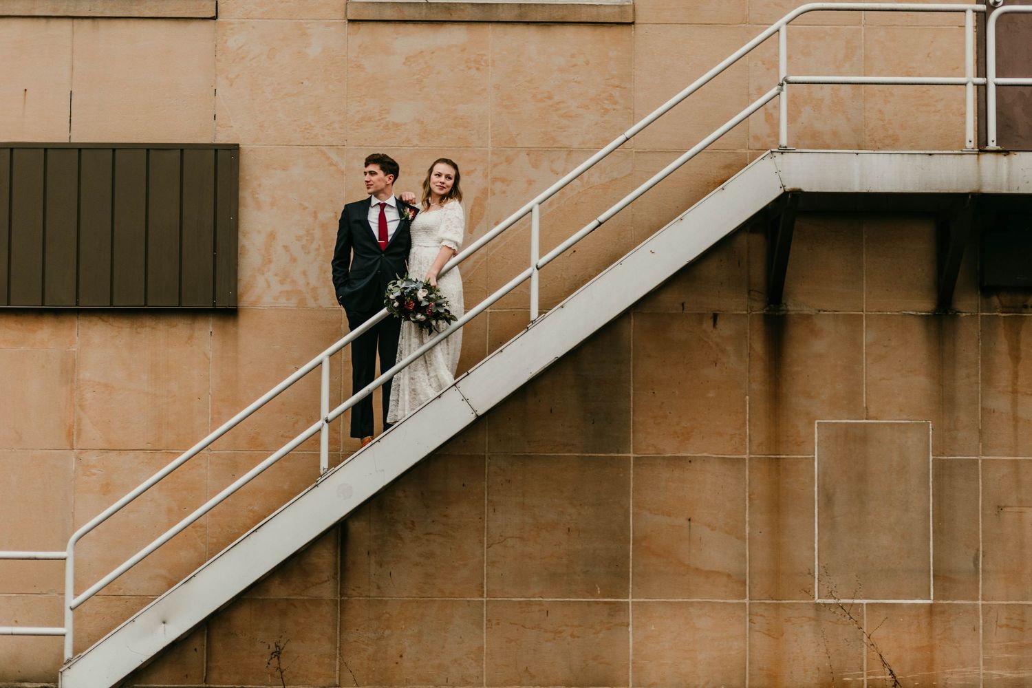 Bride and groom posing on industrial metal staircase against a stone building wall.