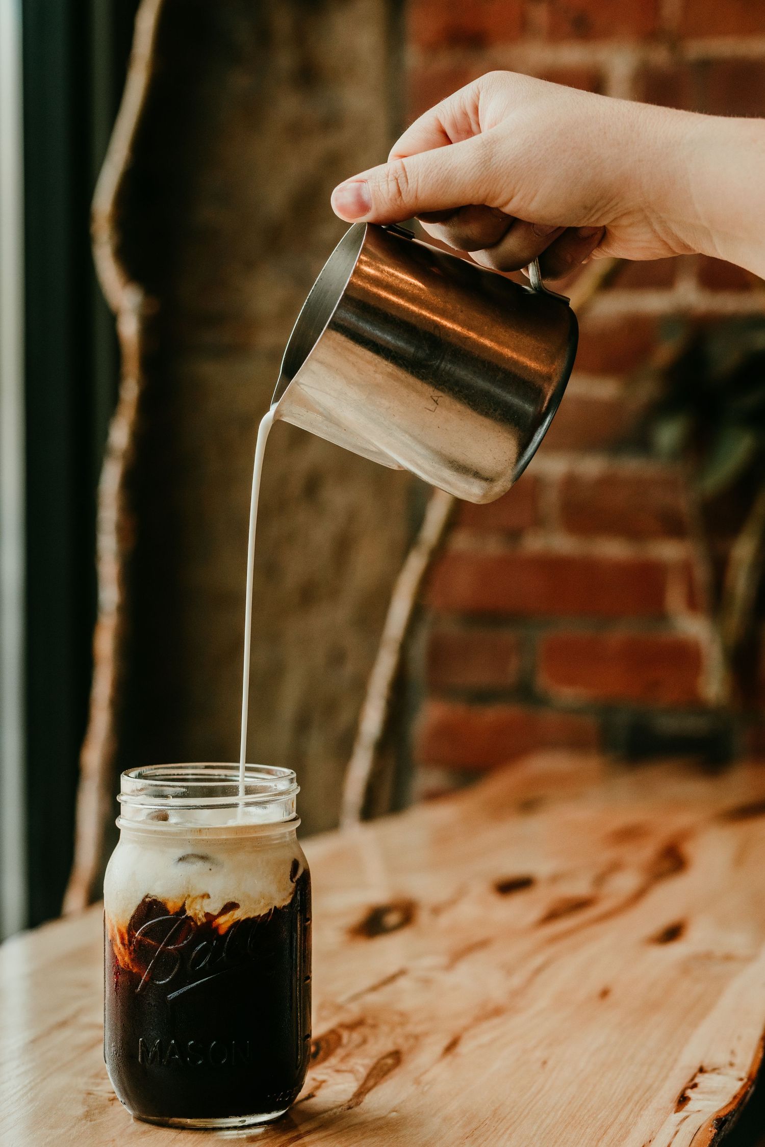Hand pouring creamy milk into iced coffee in a mason jar on a rustic wooden table.