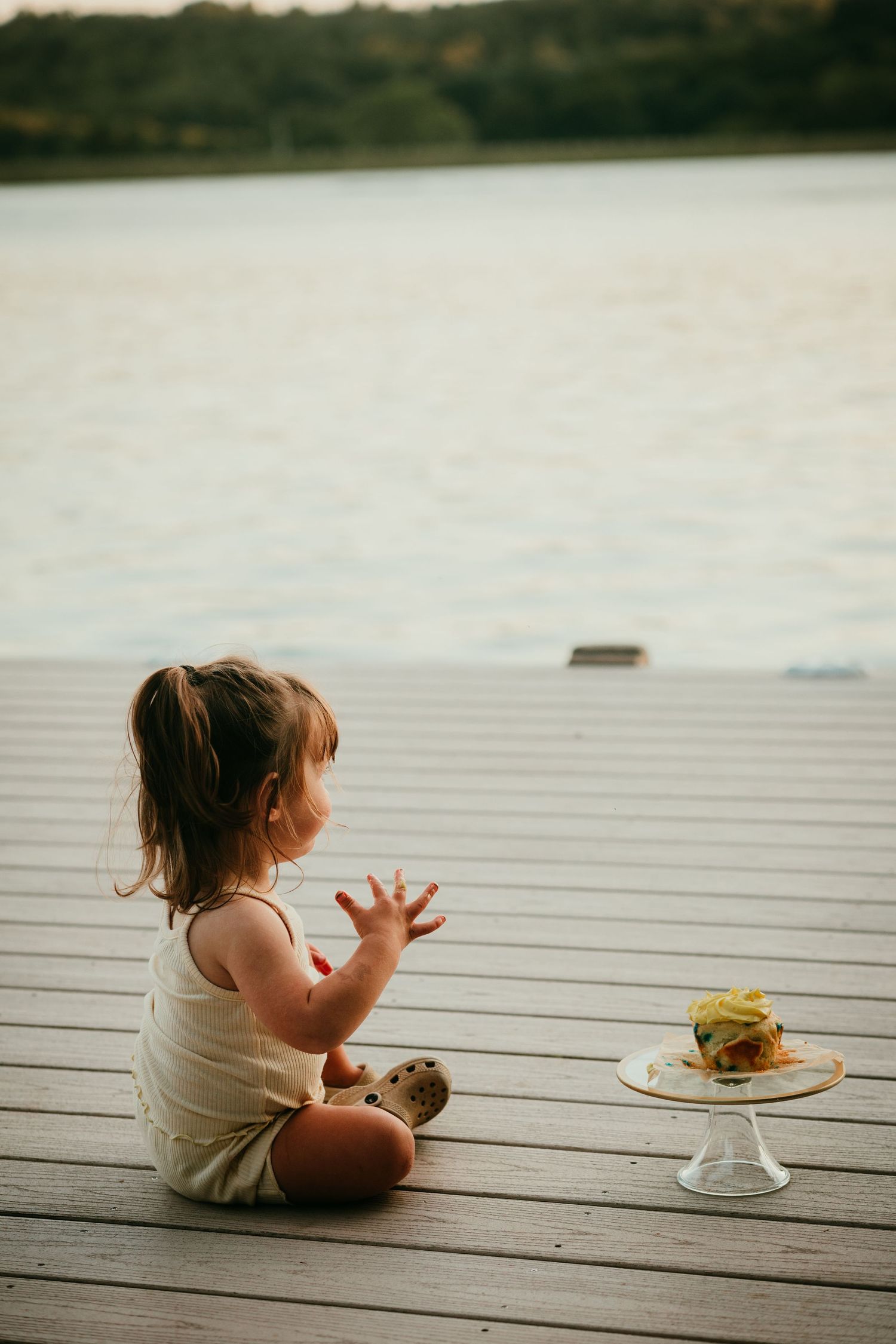 Toddler girl sitting on a wooden dock reaching toward a small cake with a lake in the background.
