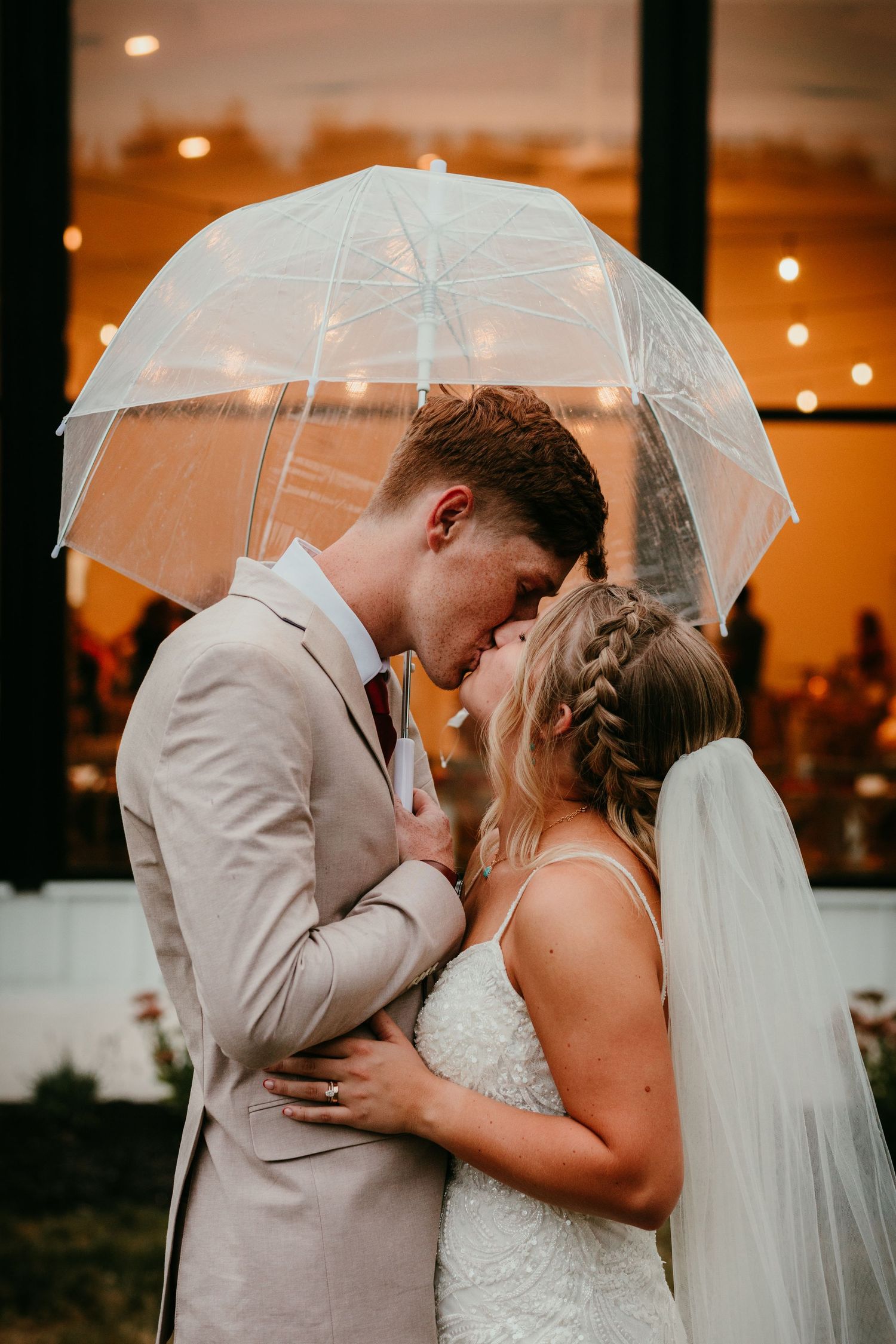 Bride and groom sharing a romantic kiss under a clear umbrella in the rain on their wedding day.