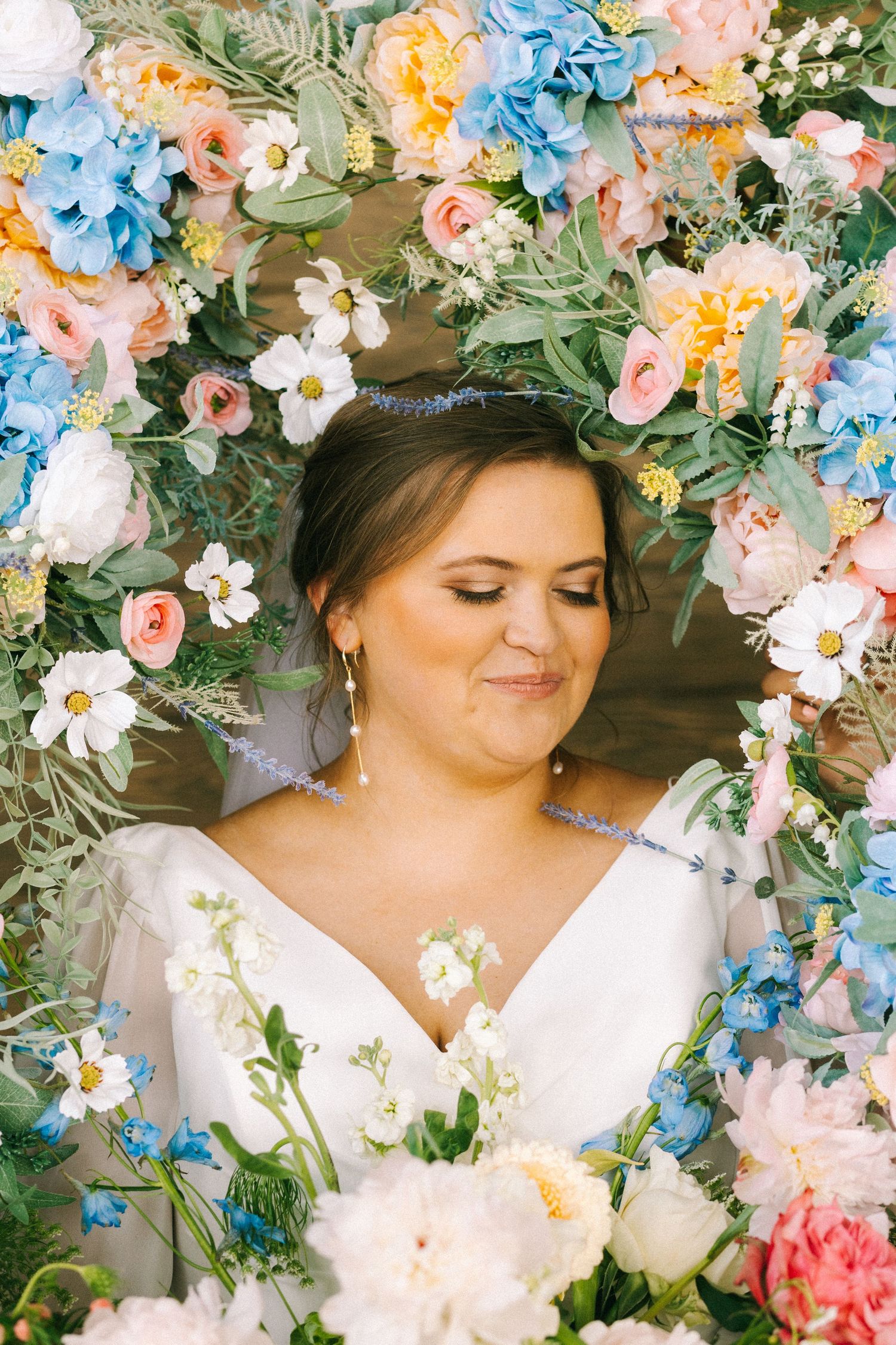 Bride smiling serenely surrounded by a stunning floral arch of blue, pink, and white wedding flowers.