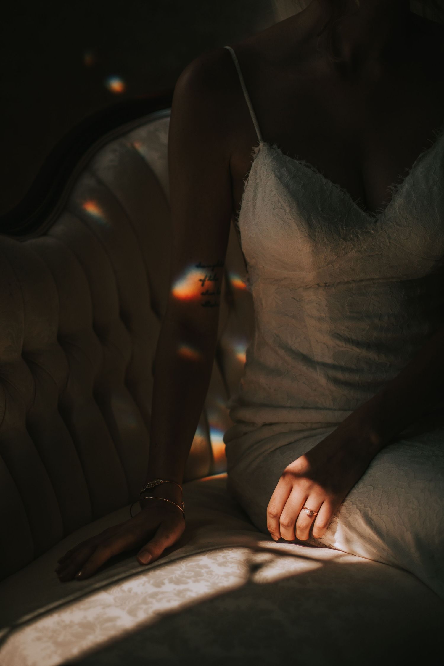 Bride in white lace wedding dress sitting in chair with sunlight and lens flare casting warm shadows.