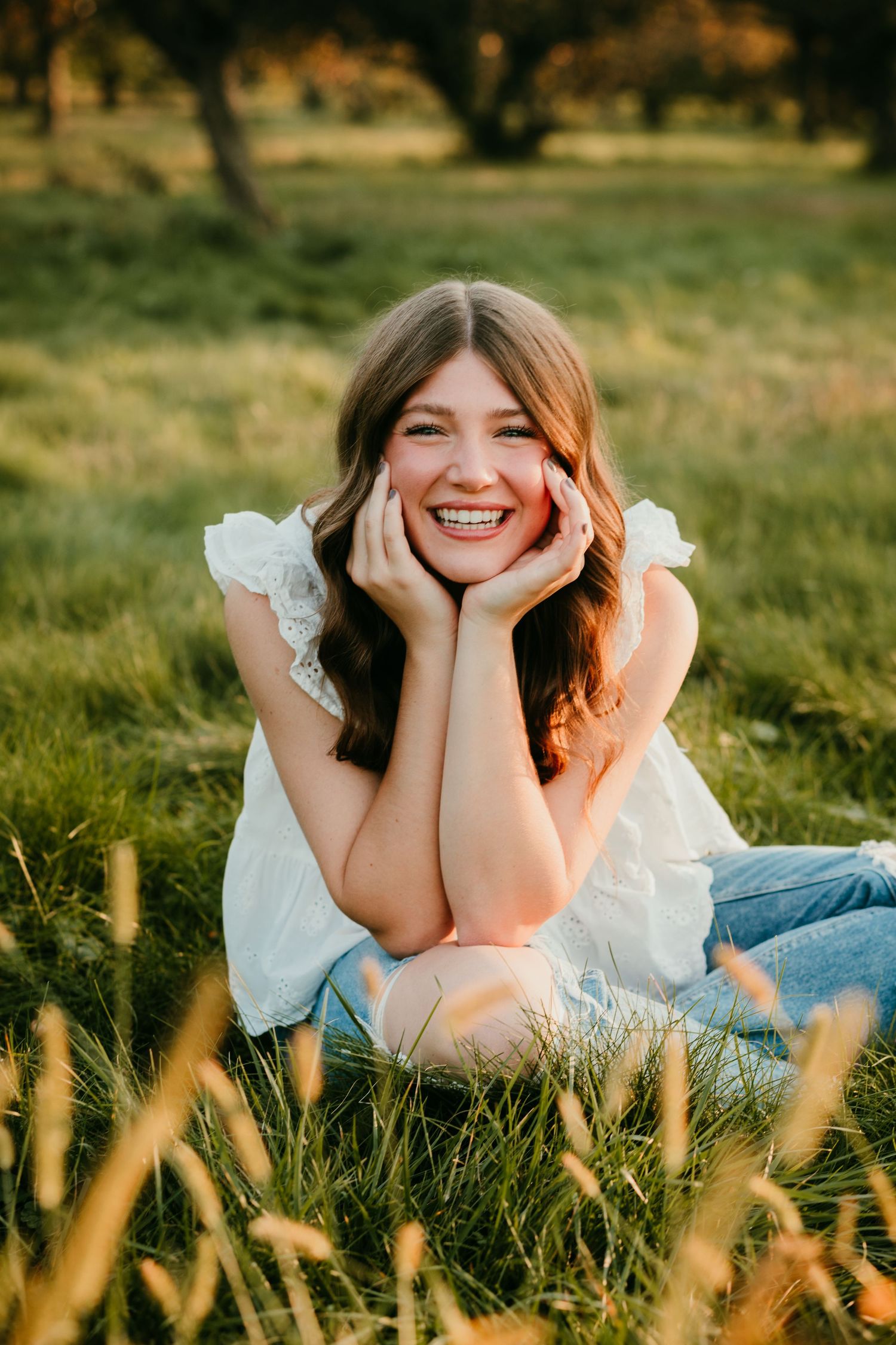Happy teenage girl with brown hair lying in green grass smiling with hands on her cheeks at sunset.