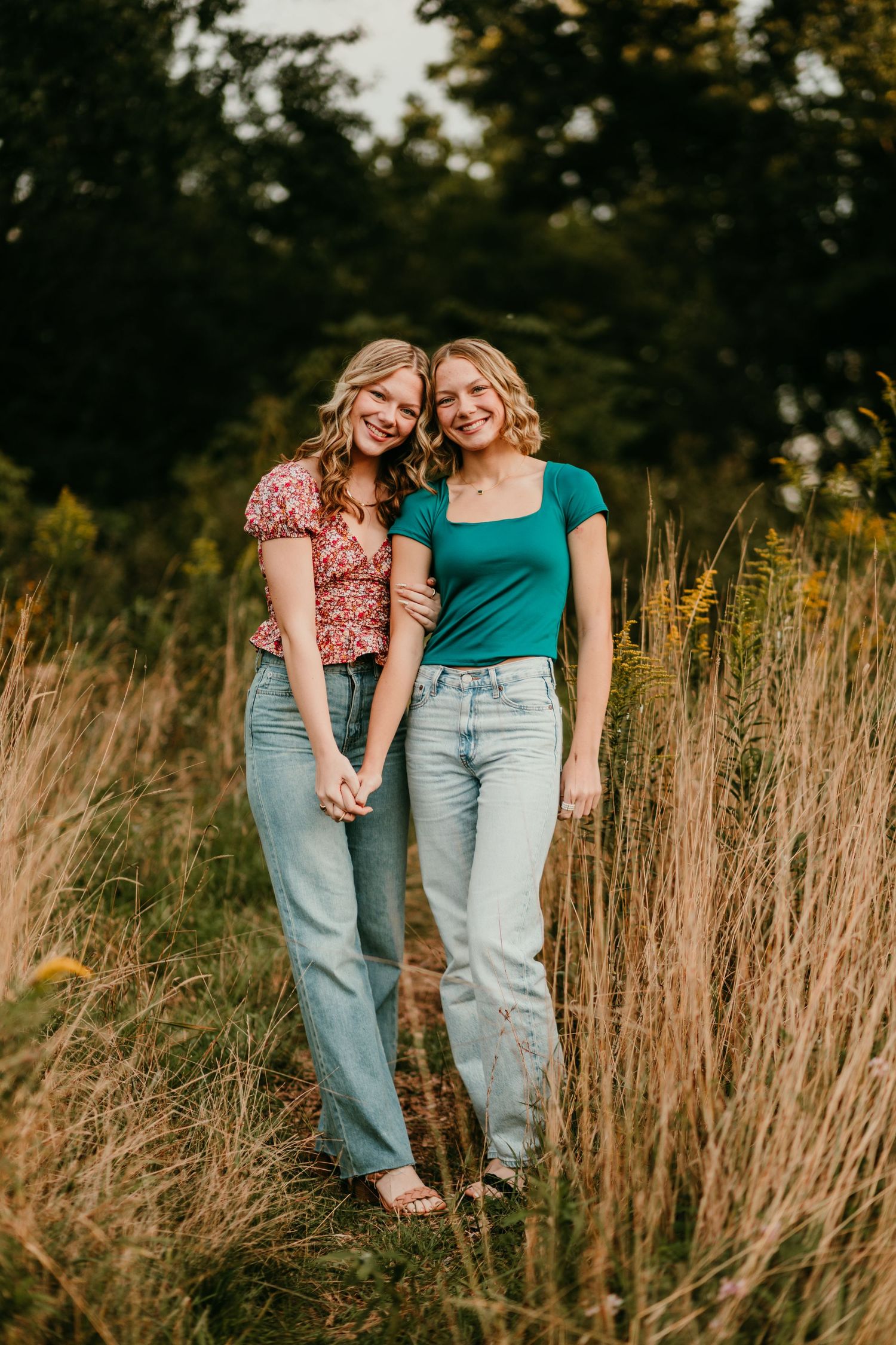 Two smiling young women holding hands in a sunlit meadow wearing casual tops and light-wash jeans.